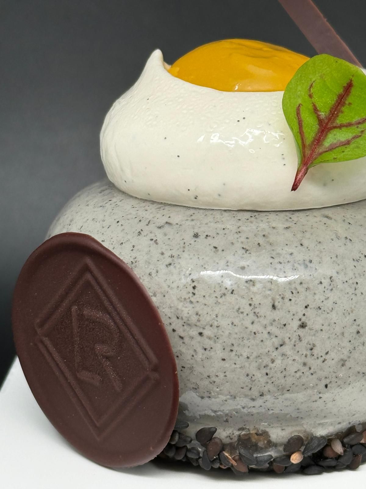Dessert avec un glaçage moucheté gris, garni de meringue blanche et d'une sauce jaune, d'un emblème en chocolat et d'une feuille verte.