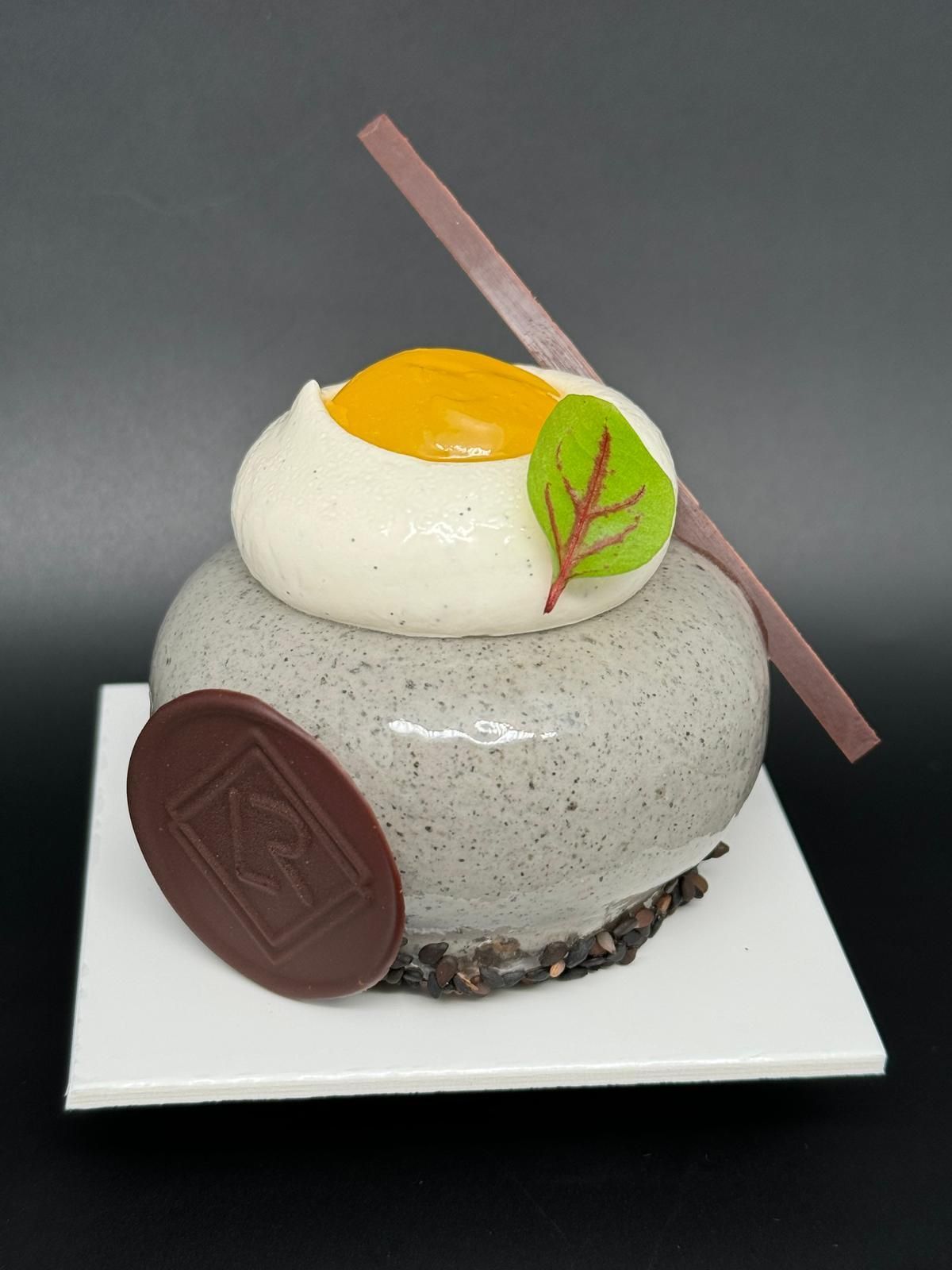 Gâteau gourmand avec glaçage gris, dessus blanc avec centre jaune, accents de chocolat et petite feuille.