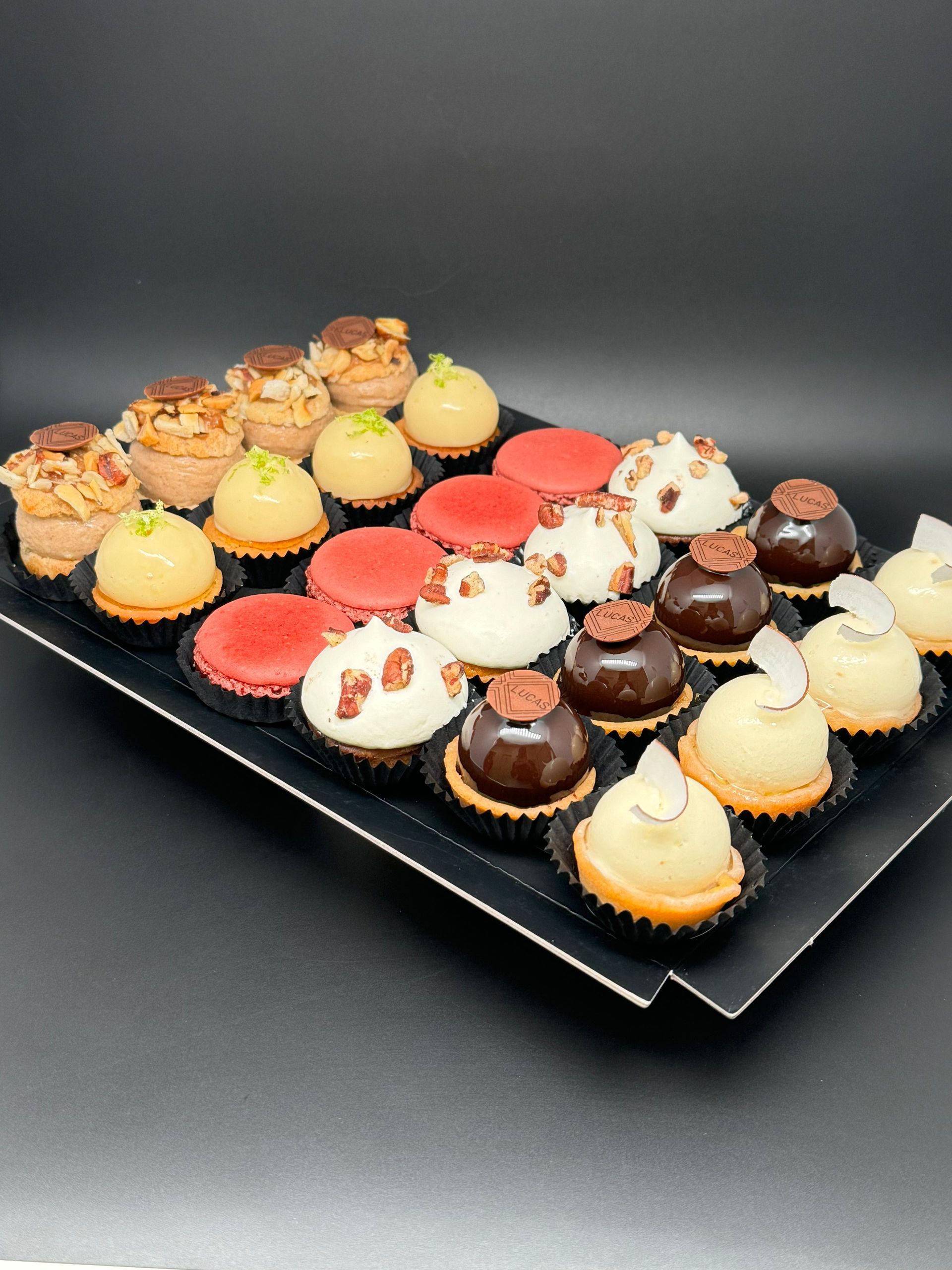 Plateau de pâtisseries miniatures assorties sur une surface noire; certaines sont rondes, d'autres garnies de noix, de chocolat ou de glaçage.