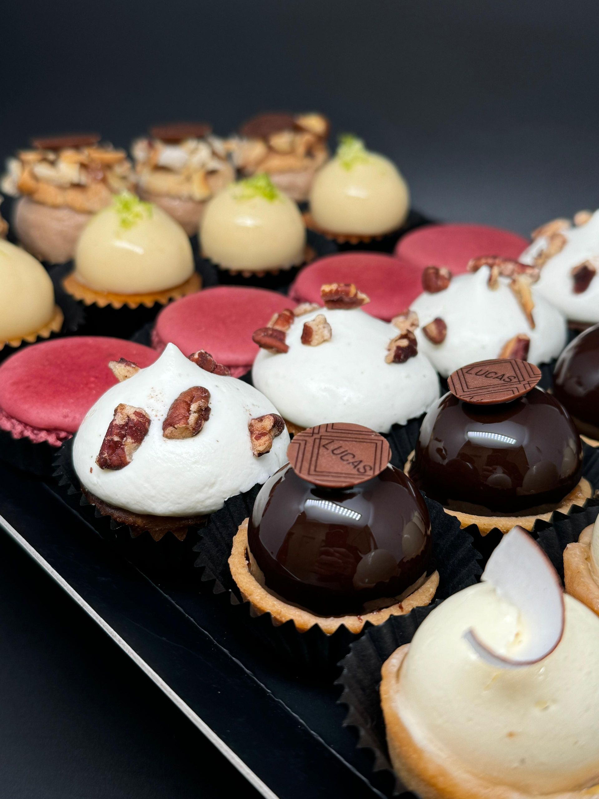 Plateau de mini pâtisseries assorties, comprenant des tartelettes, des macarons et des desserts garnis de crème, sur fond noir.