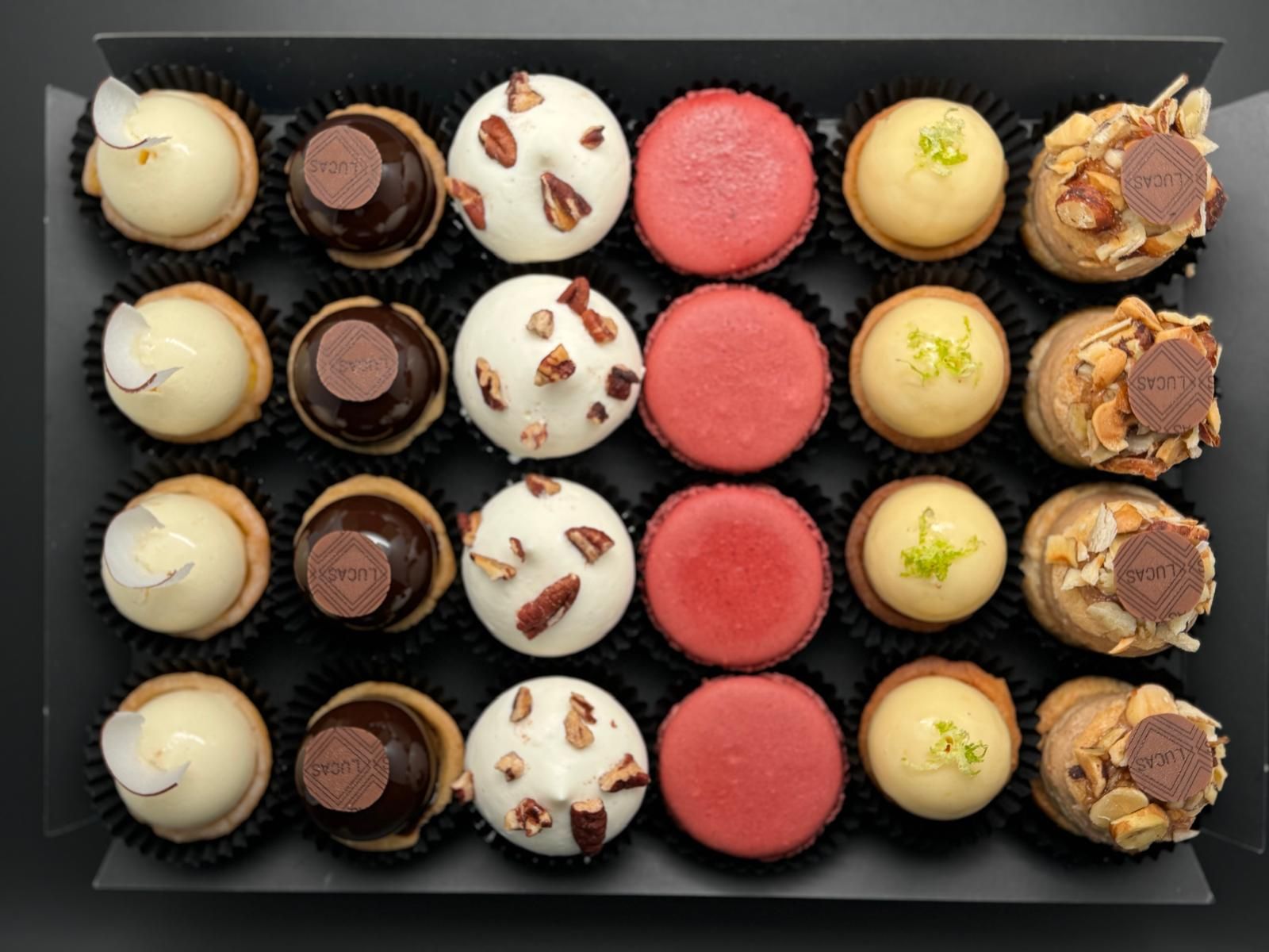 Assortiment de pâtisseries gourmandes dans une boîte noire : boules de noix de coco, tartelettes au chocolat, gourmandises garnies de pacanes, macarons rouges, desserts au citron vert et rouleaux de noix.