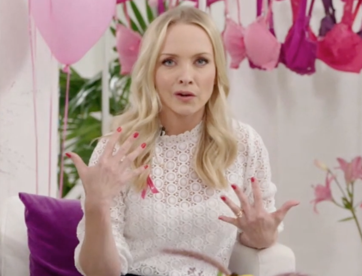 Eine Frau in einem weißen Spitzentop sitzt auf einem Sofa. Im Hintergrund sind rosa Luftballons zu sehen.