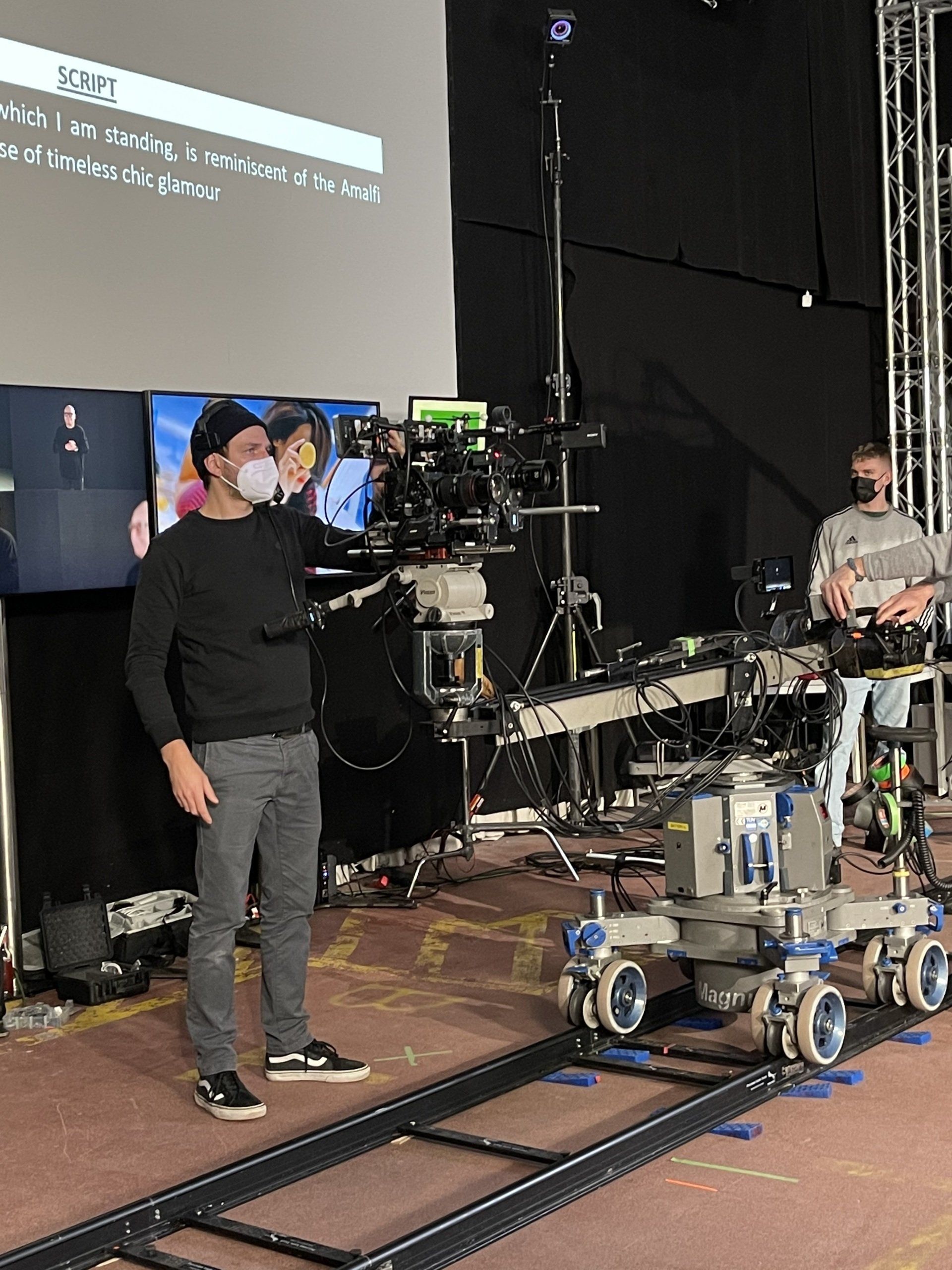 Ein Mann mit einer Maske steht auf einer Rollbahn vor einem großen Bildschirm.