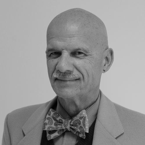 Bald man with mustache, wearing a patterned bow tie and suit jacket, smiles in front of a white background.