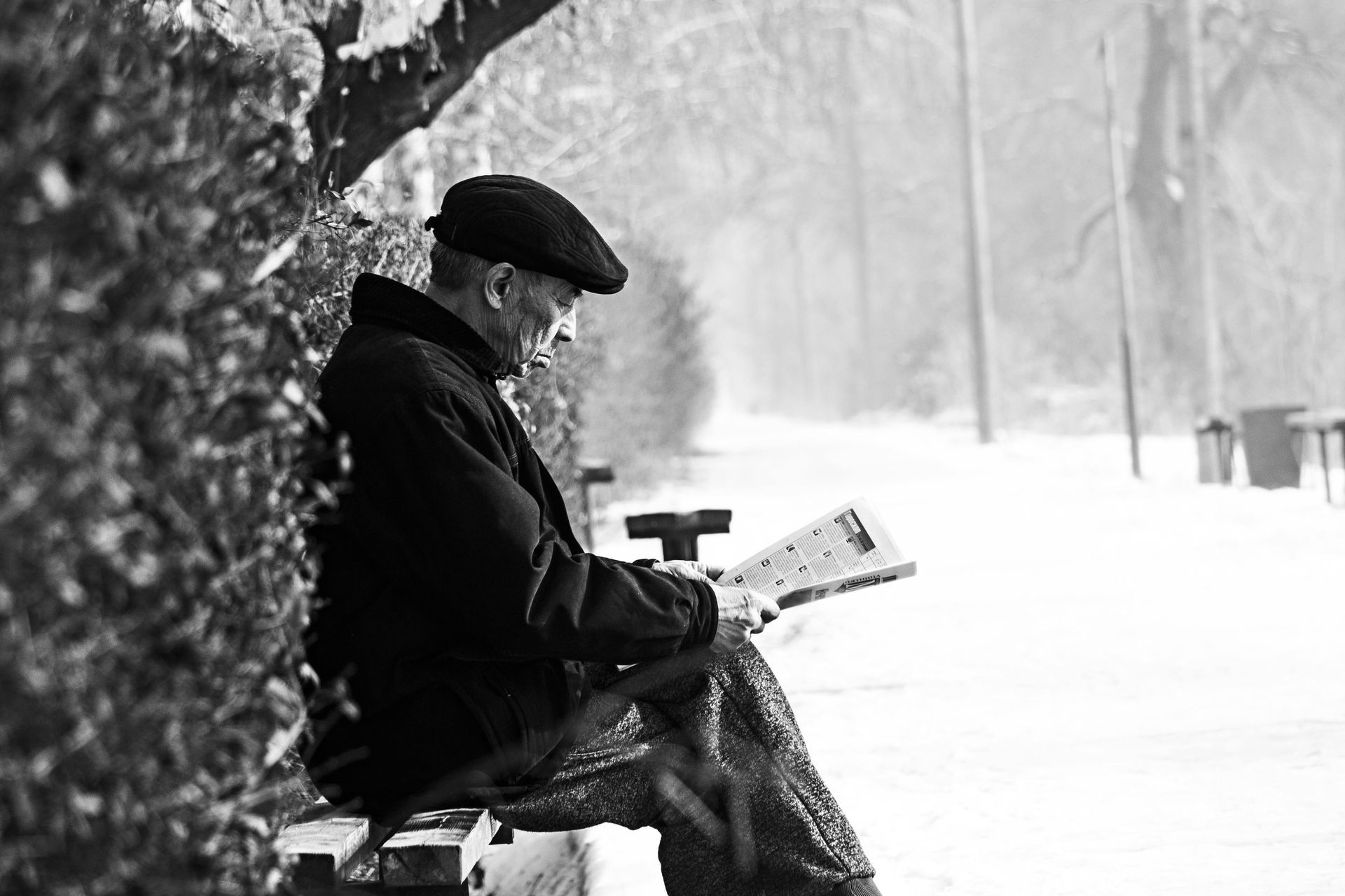 An elderly man is sitting on a park bench reading a newspaper.