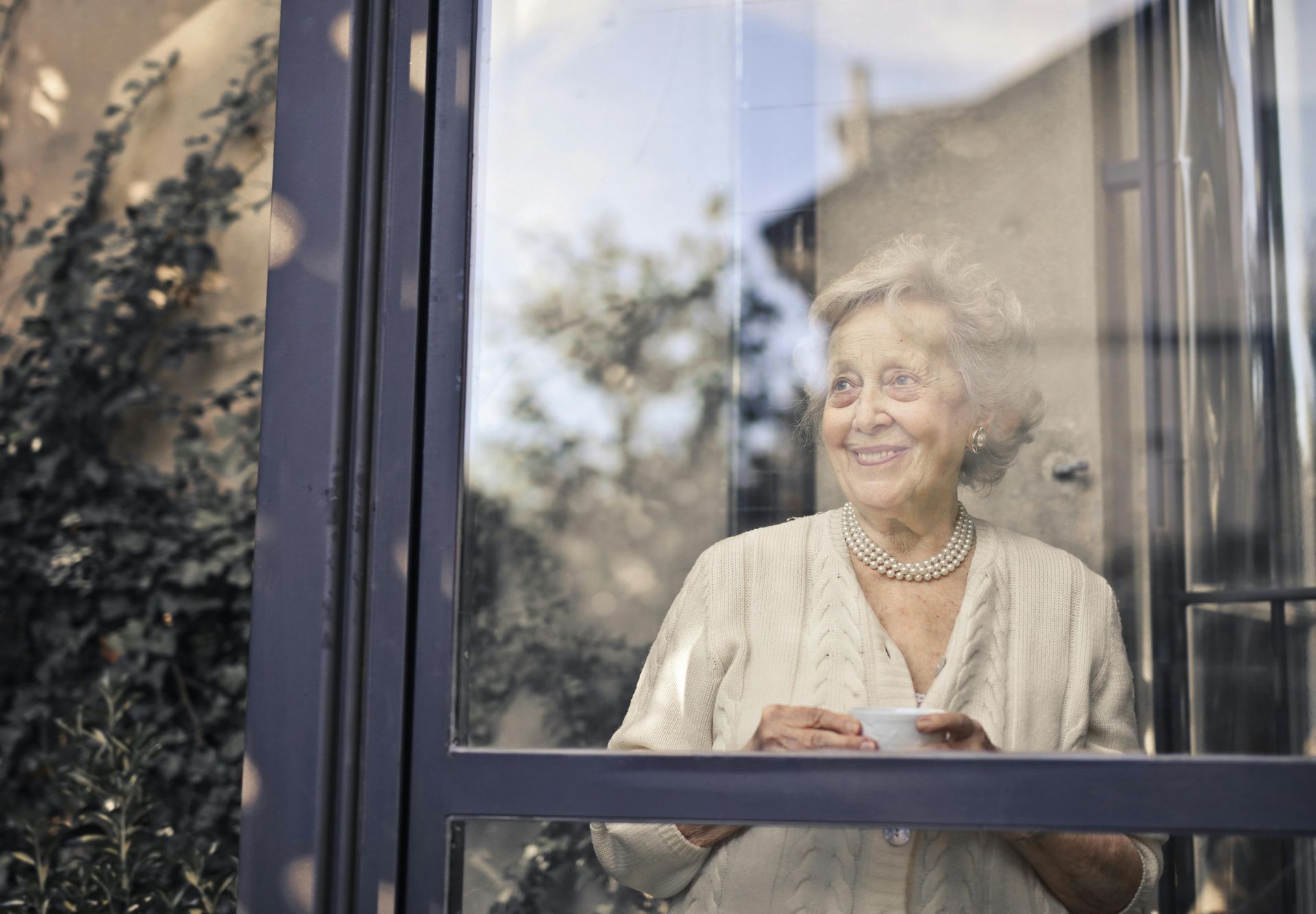An elderly woman is looking out of a window while holding a cup of coffee.