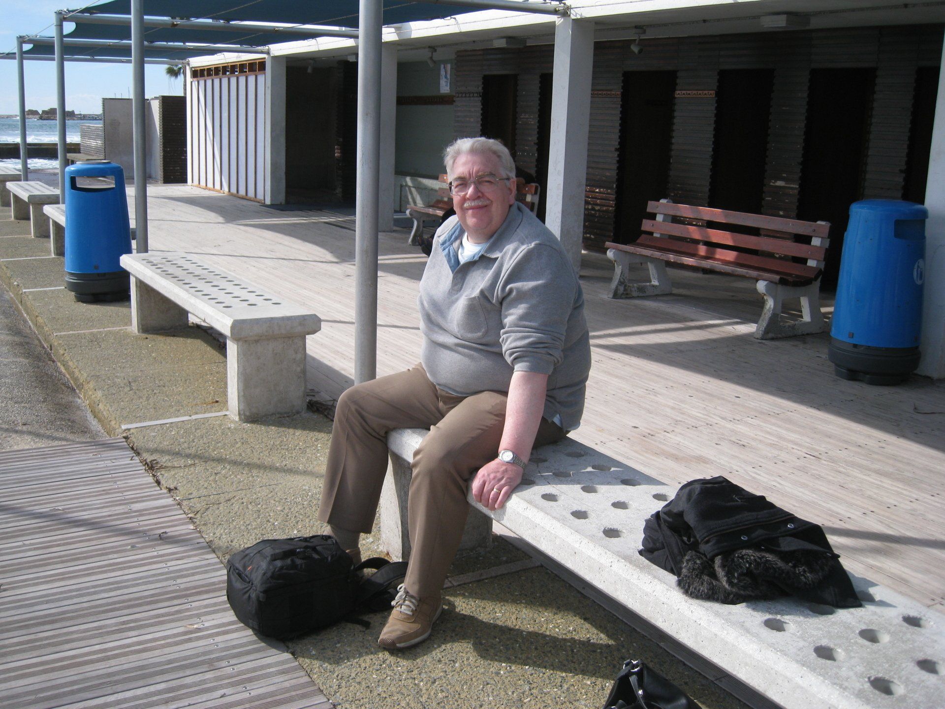A man sits on a bench in front of a blue trash can