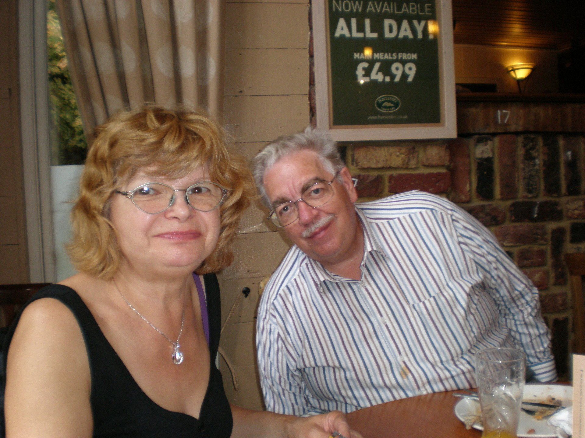 A man and woman sit at a table in front of a sign that says now available all day