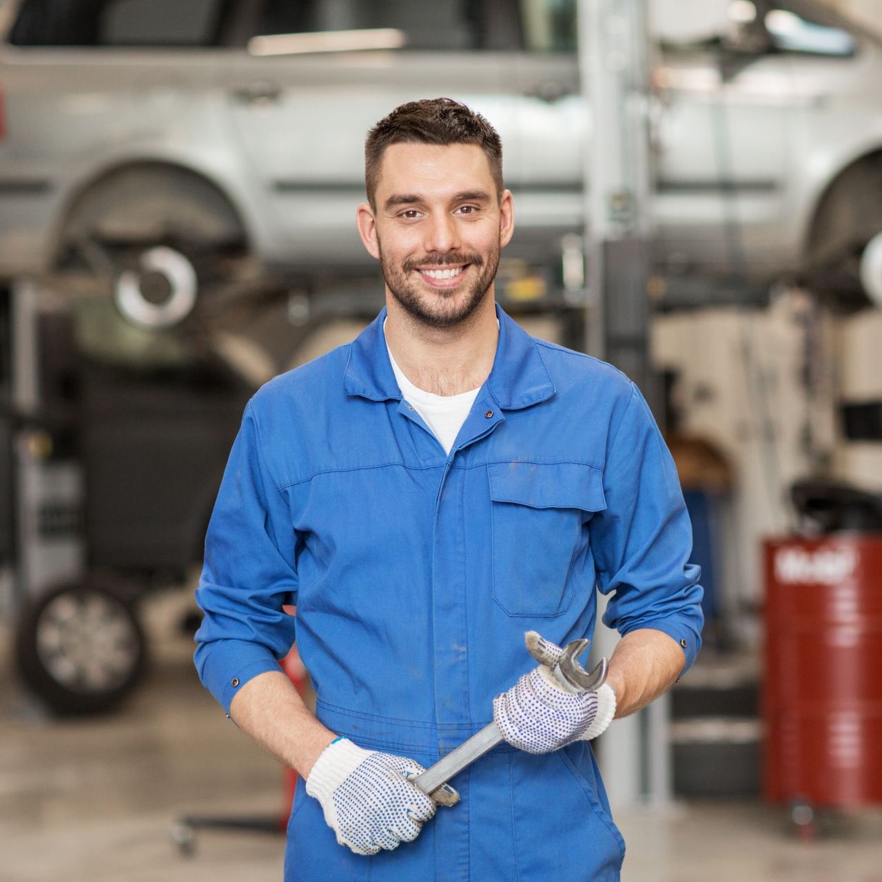 Mechanic in blue overalls smiles, holding a wrench in a garage with a car.