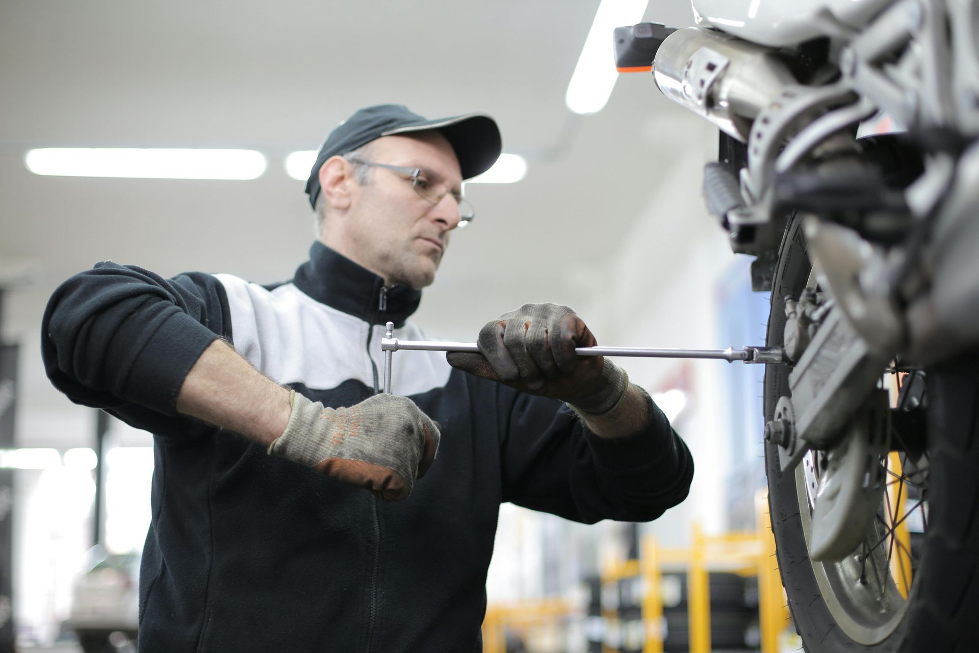 Mechanic in a shop repairing a motorcycle wheel with a wrench. Wearing gloves, cap, glasses, and a black and white jacket.