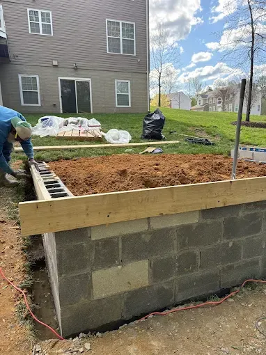 Workers building a raised cinder block foundation beside a house, with dirt fill and wooden form boards.