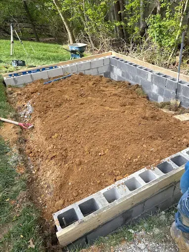 Partially built cinder block foundation with dirt fill in a wooded backyard