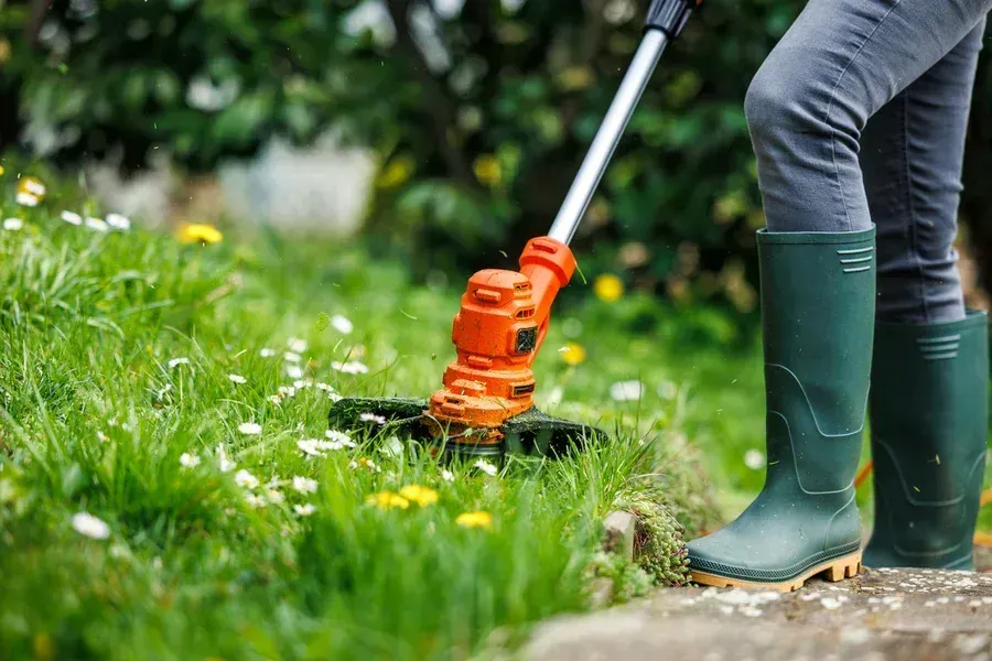 Person trimming grass with a string trimmer beside a lawn, wearing green boots.