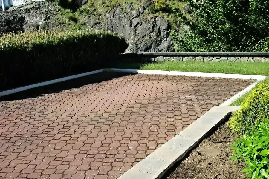 Empty brick-paved parking area beside a rocky hillside and green hedge in bright sunlight
