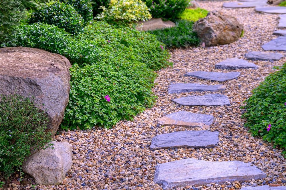 Stepping stone path through a green garden with rocks and pink flowers, bordered by gravel.