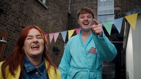 Two people laughing outdoors beside colorful bunting, with one pointing toward the camera.