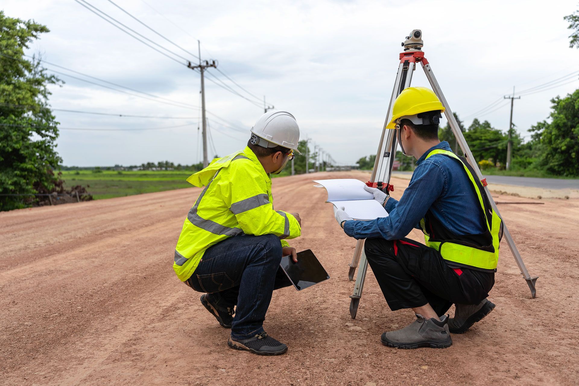 Engineer holding tablet to inspect work, with foreman using telescope to check pin against road plan, telescope and radio to control road construction, against road background under construction