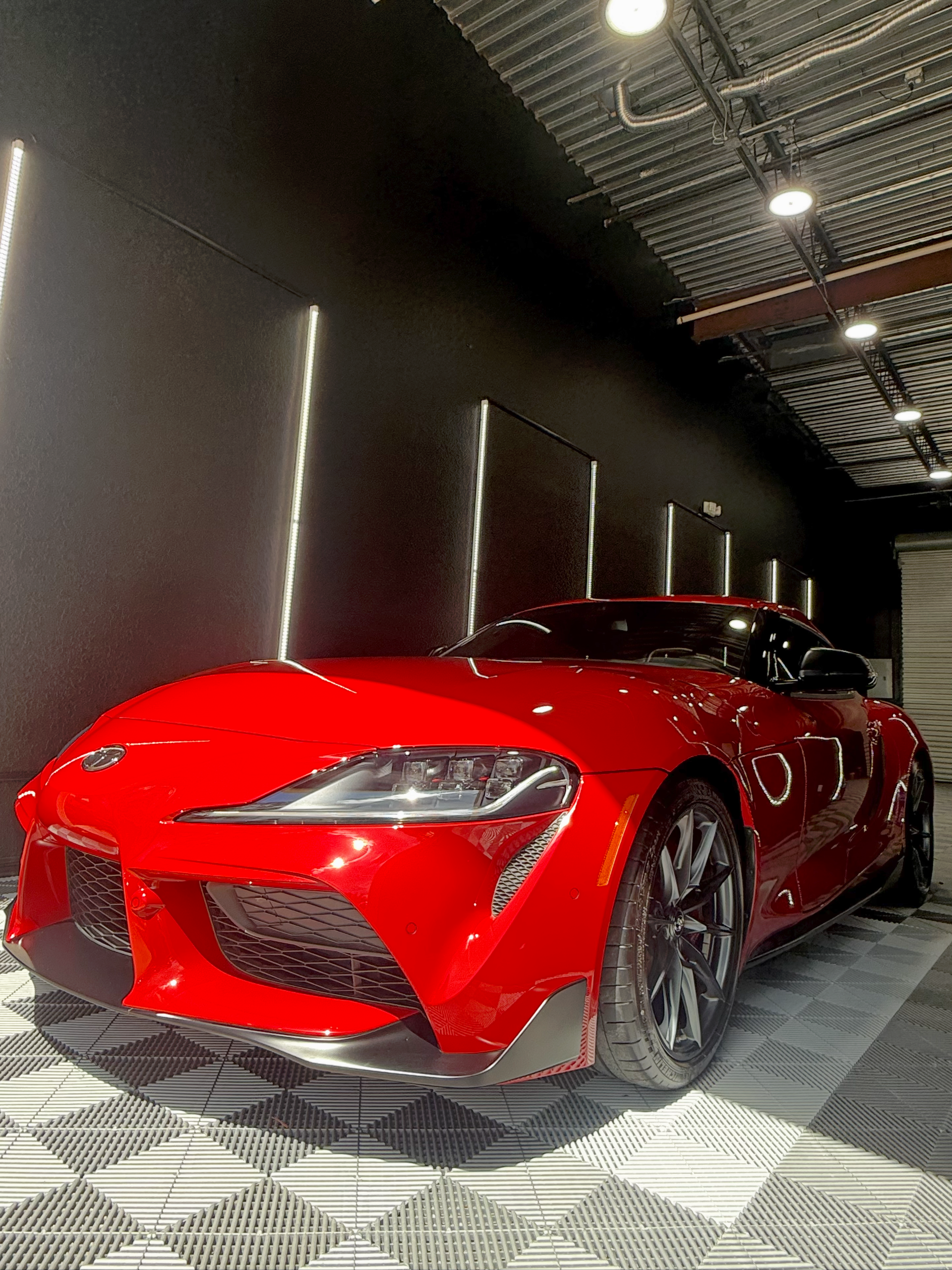 Red Toyota Supra sports car parked in a garage with black walls, featuring neon lights.