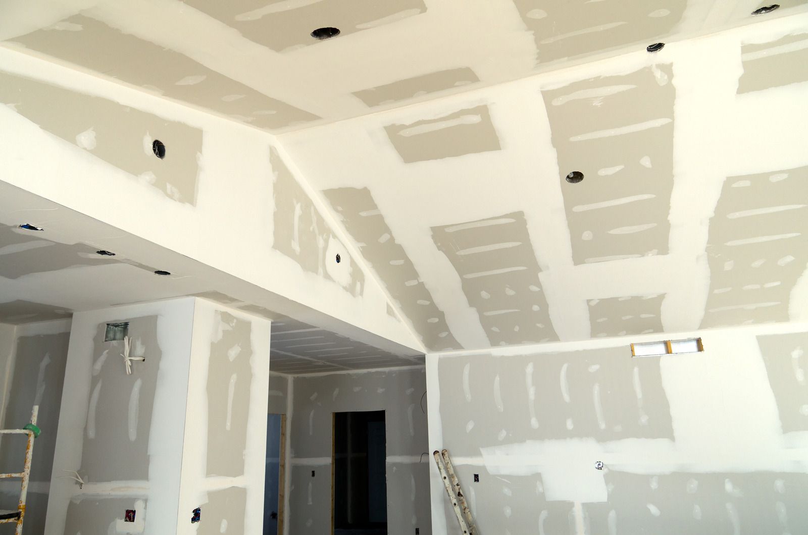 Interior of a room under construction, with drywall covering walls and ceiling; recessed lighting fixtures are visible.
