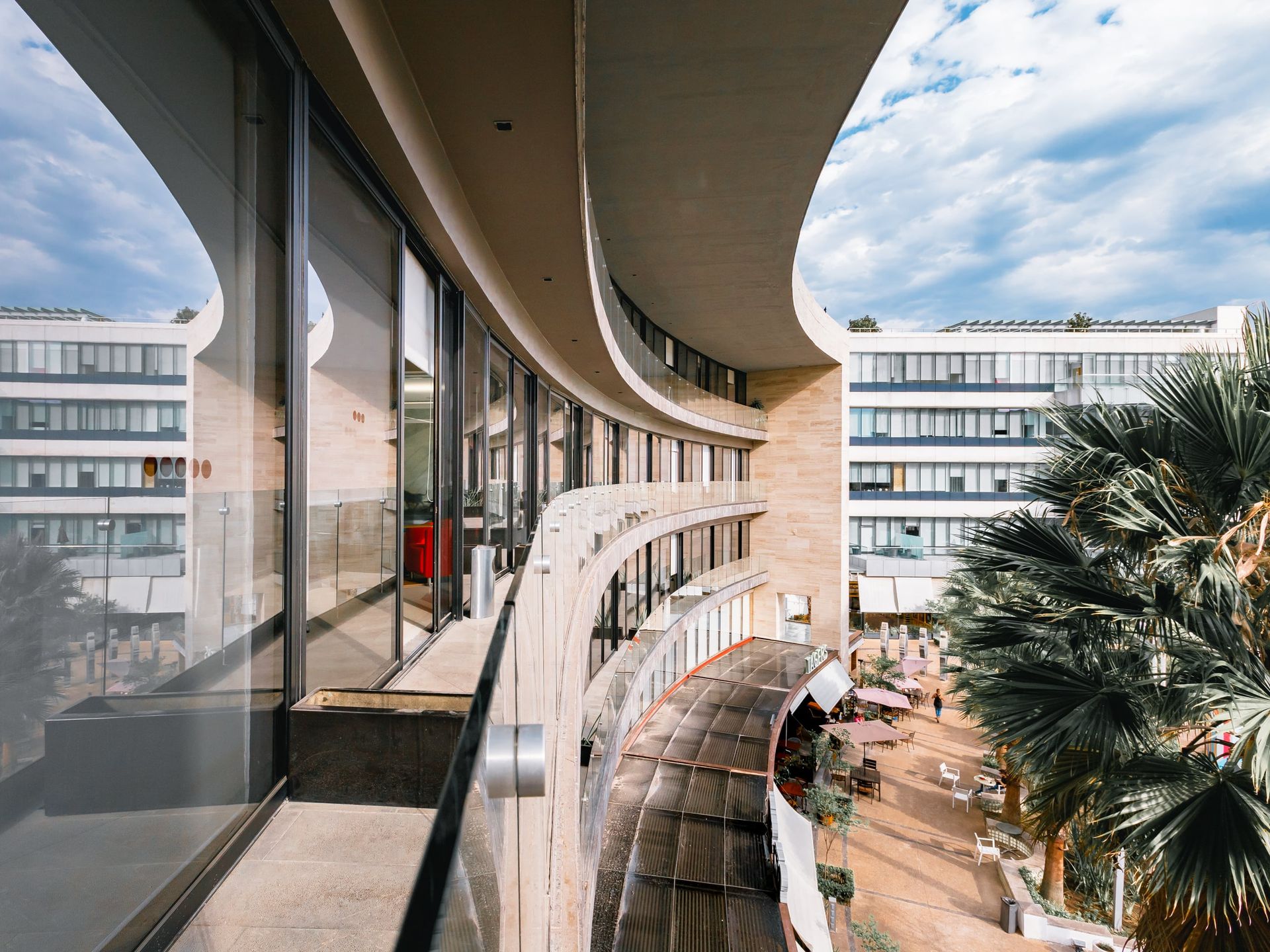 Edificio de hormigón curvo con grandes ventanales y balcón con vista a un patio con palmeras.