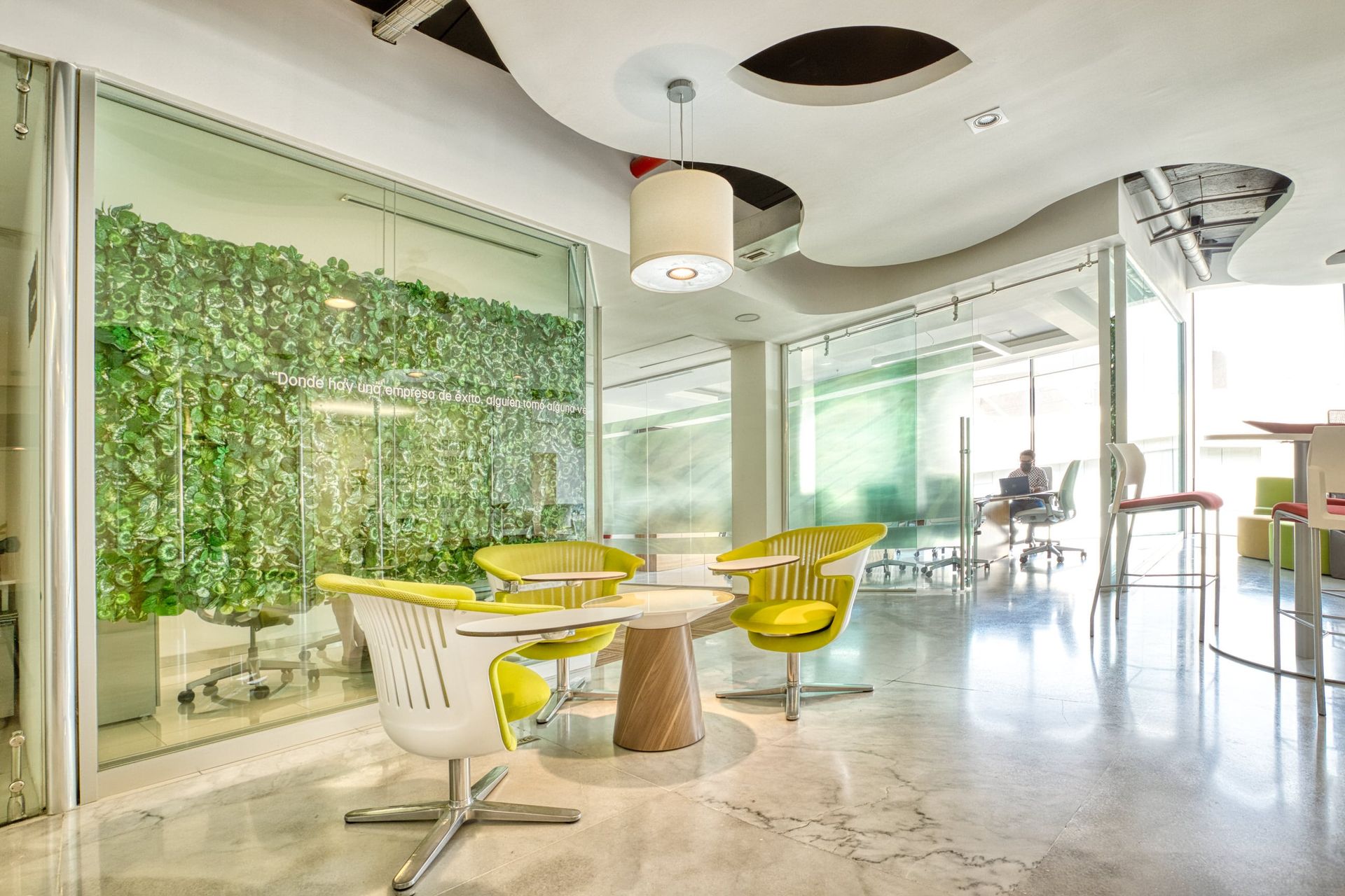 Espacio de oficina moderno con una pared viva, zona de estar con sillas amarillas y paredes de vidrio.