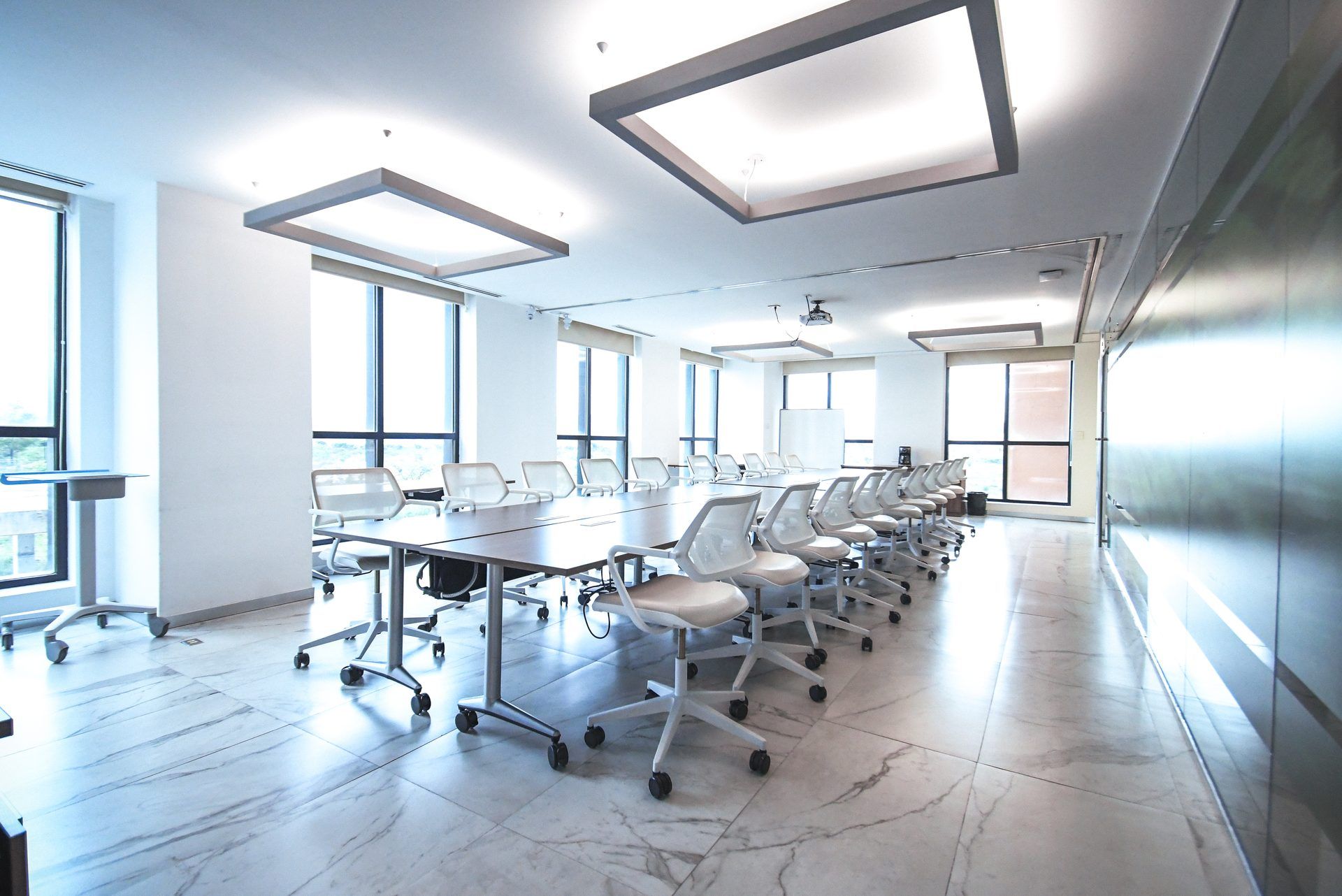 Sala de conferencias luminosa y moderna con mesas largas, sillas blancas, ventanas grandes y luces de techo rectangulares.