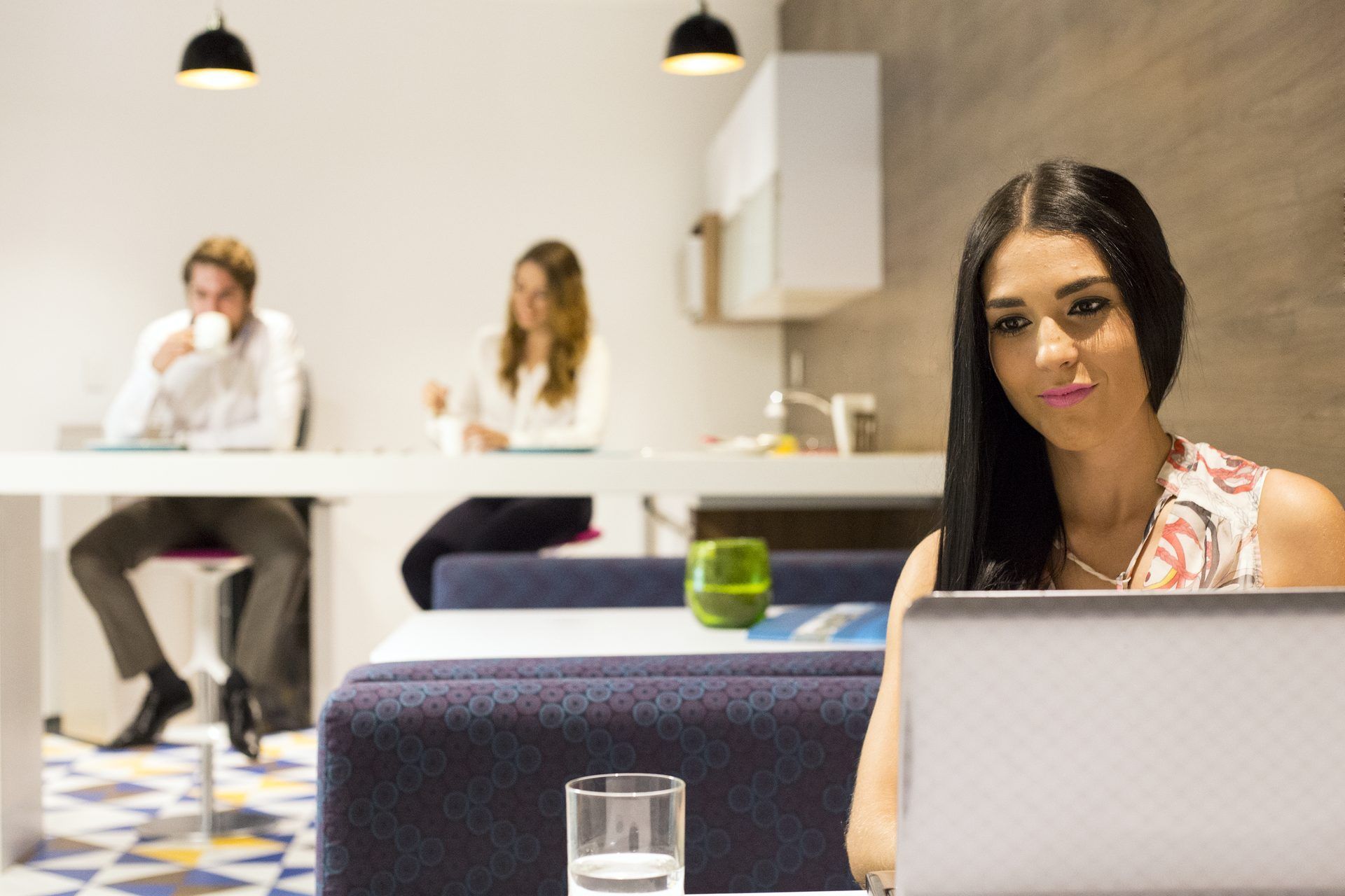 Mujer con una laptop, mirando a la cámara. Dos personas al fondo, sentadas en una mesa, en una oficina.