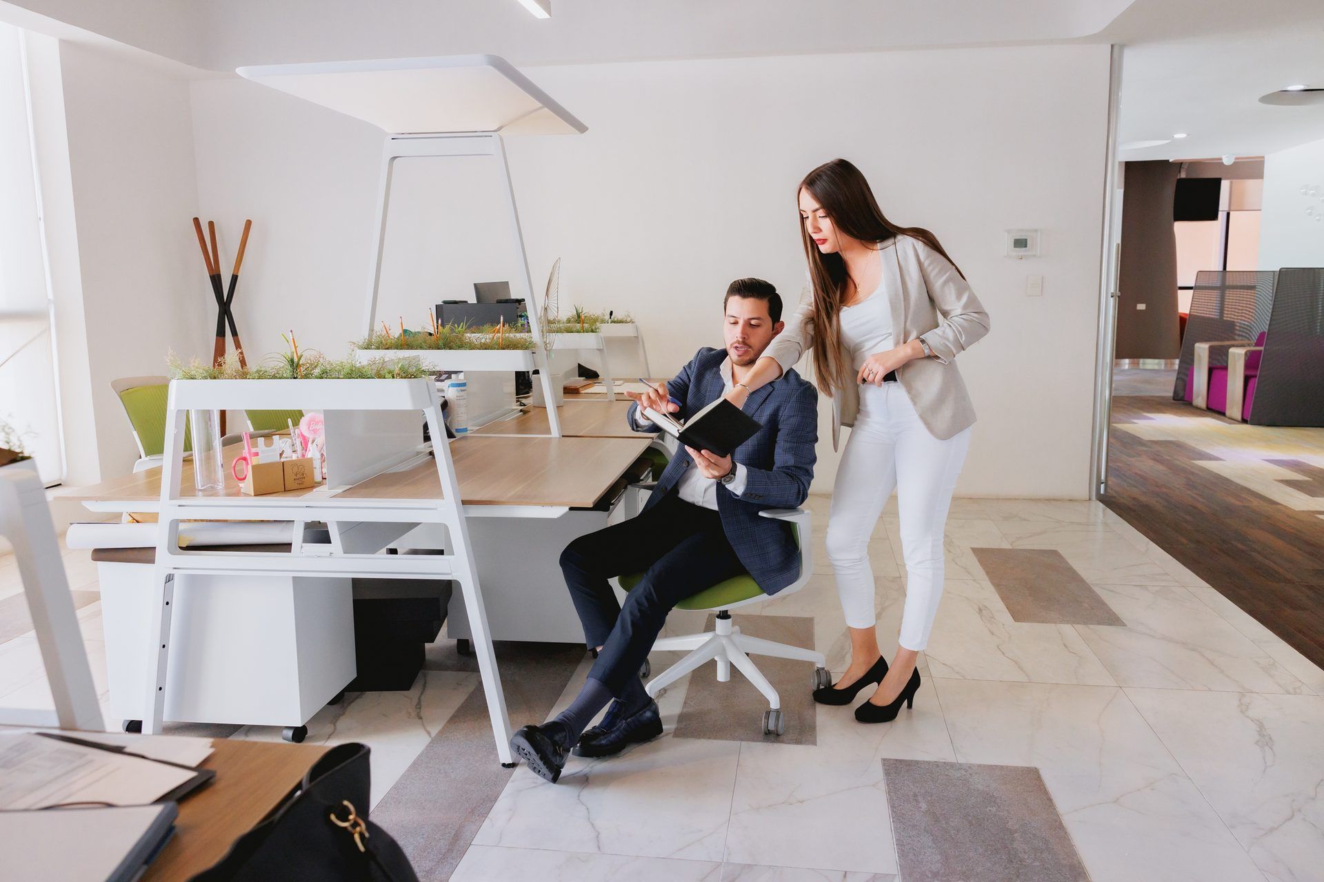 Hombre sentado en un escritorio, mirando notas; mujer de pie, señalando, en una oficina moderna.