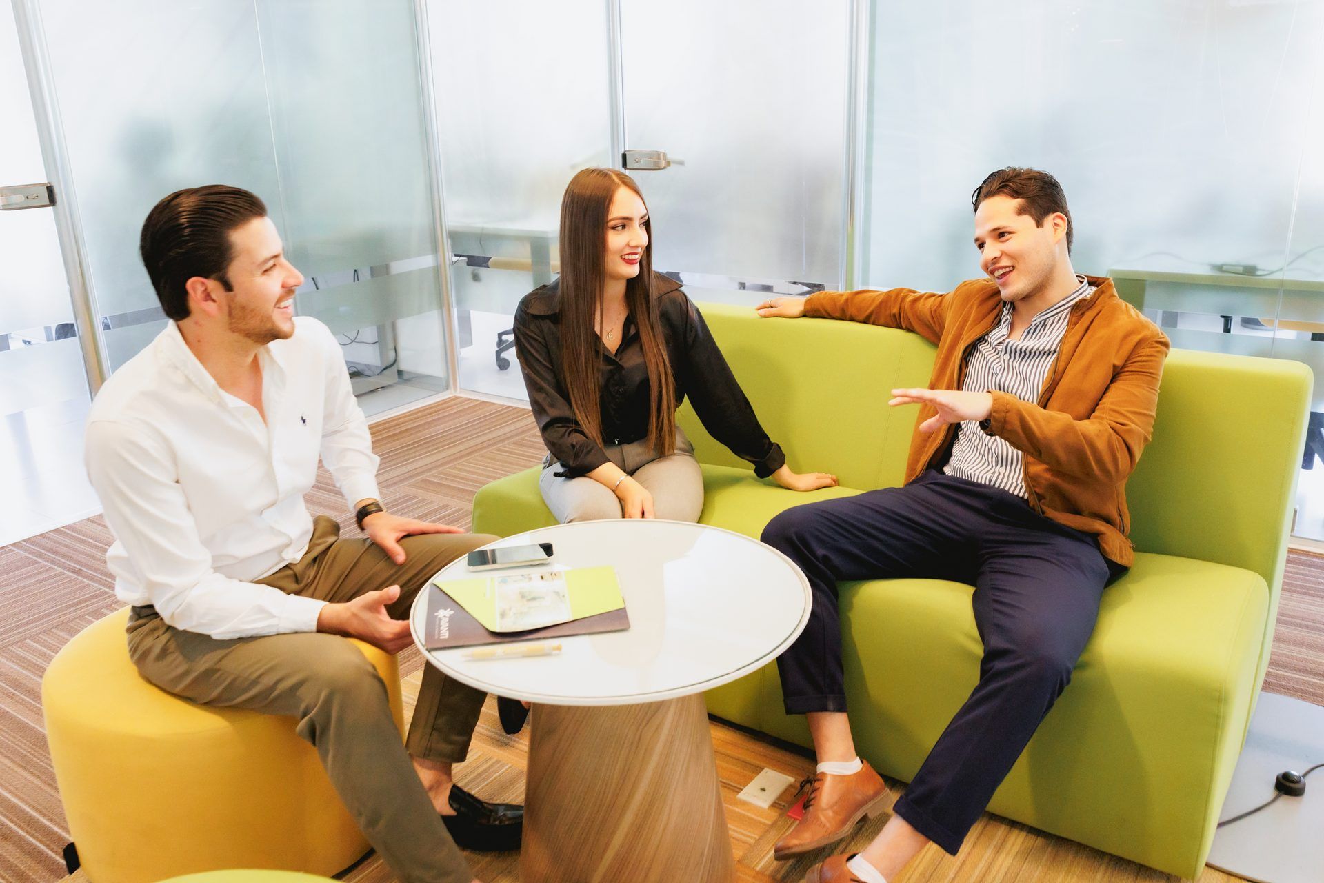 Tres personas vestidas de manera informal sentadas y conversando en un moderno salón de oficina con detalles en verde y amarillo.