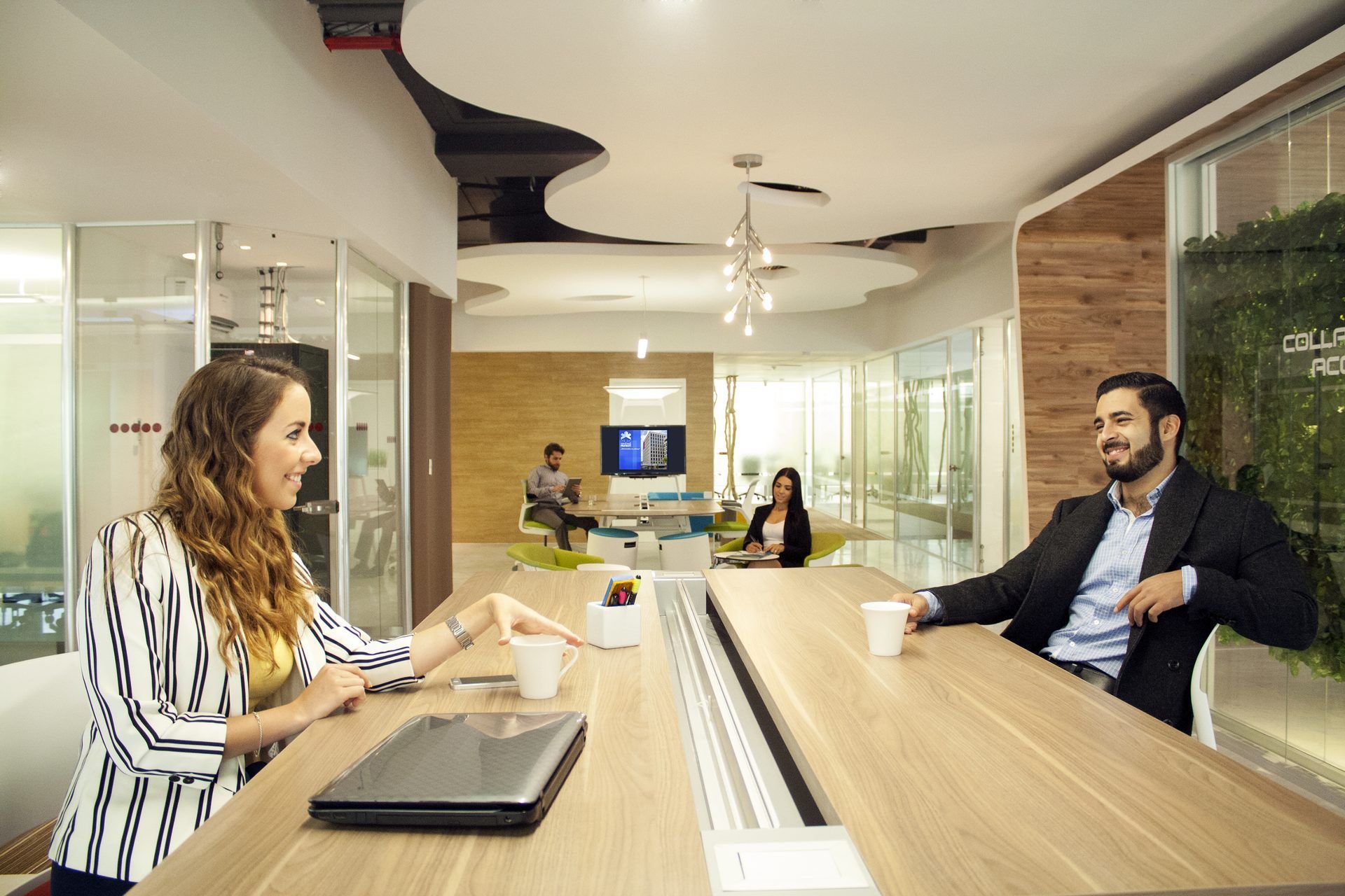Dos personas en una oficina moderna, sentadas en una mesa larga, sonriendo y conversando. Otros trabajan al fondo.