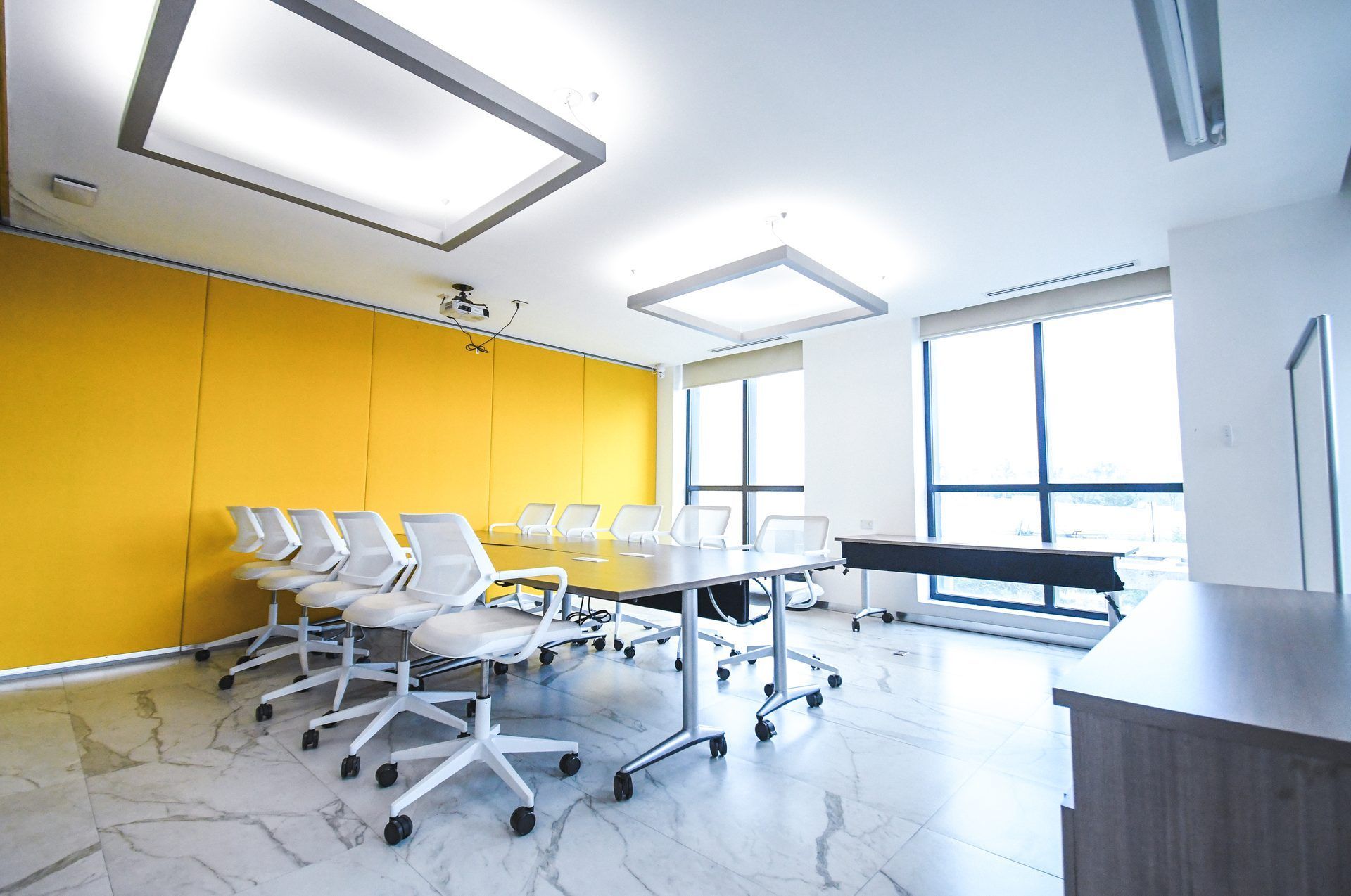 Sala de conferencias luminosa con mesa larga, sillas blancas y pared amarilla. Grandes ventanales y luces cenitales.