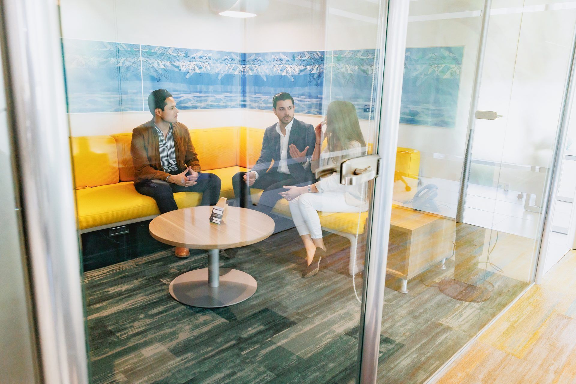 Tres personas en una reunión, sentadas en un sofá amarillo en una sala con paredes de cristal, participan en una discusión.