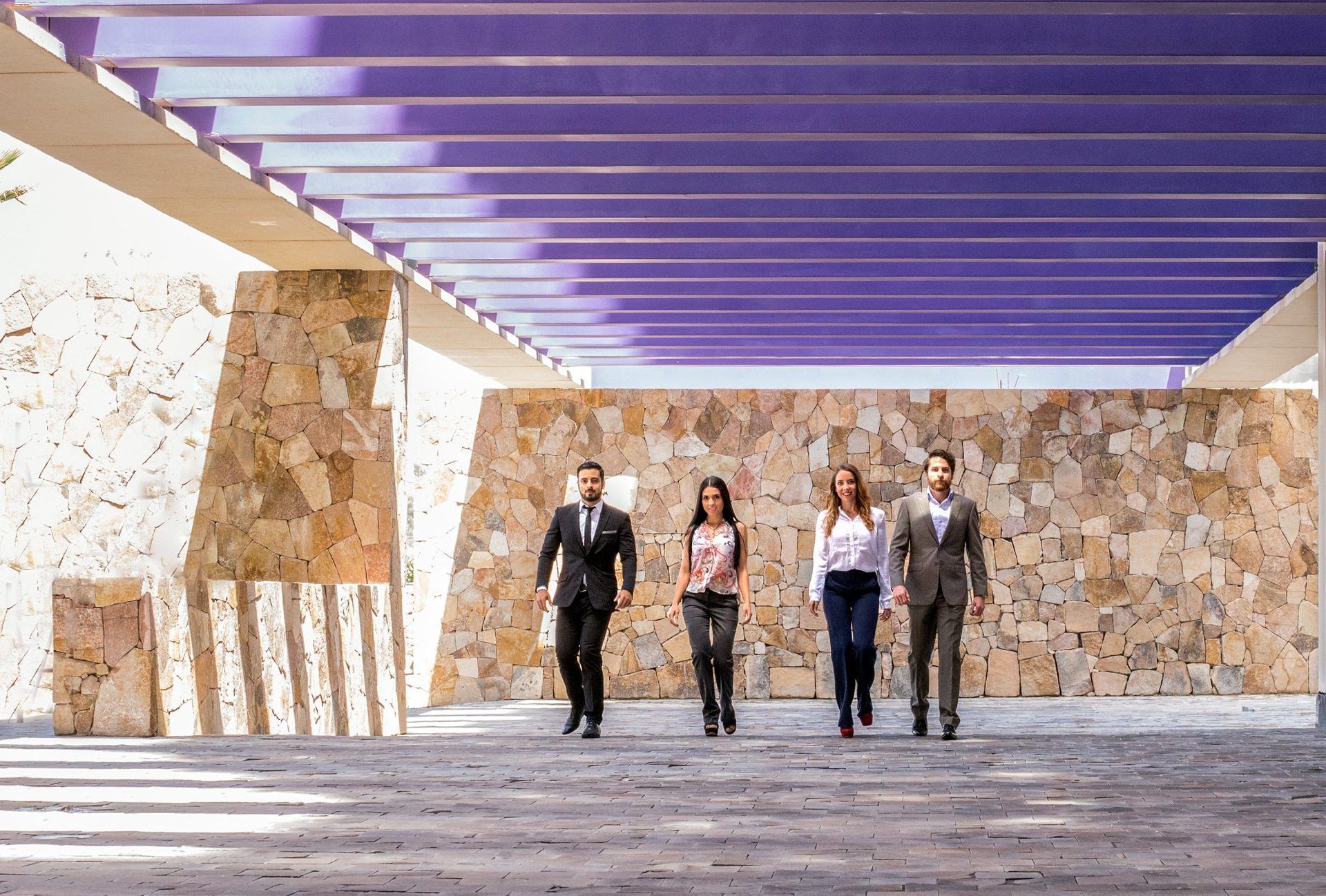 Cuatro personas caminan hacia el espectador bajo una pasarela con techo morado y paredes de piedra.
