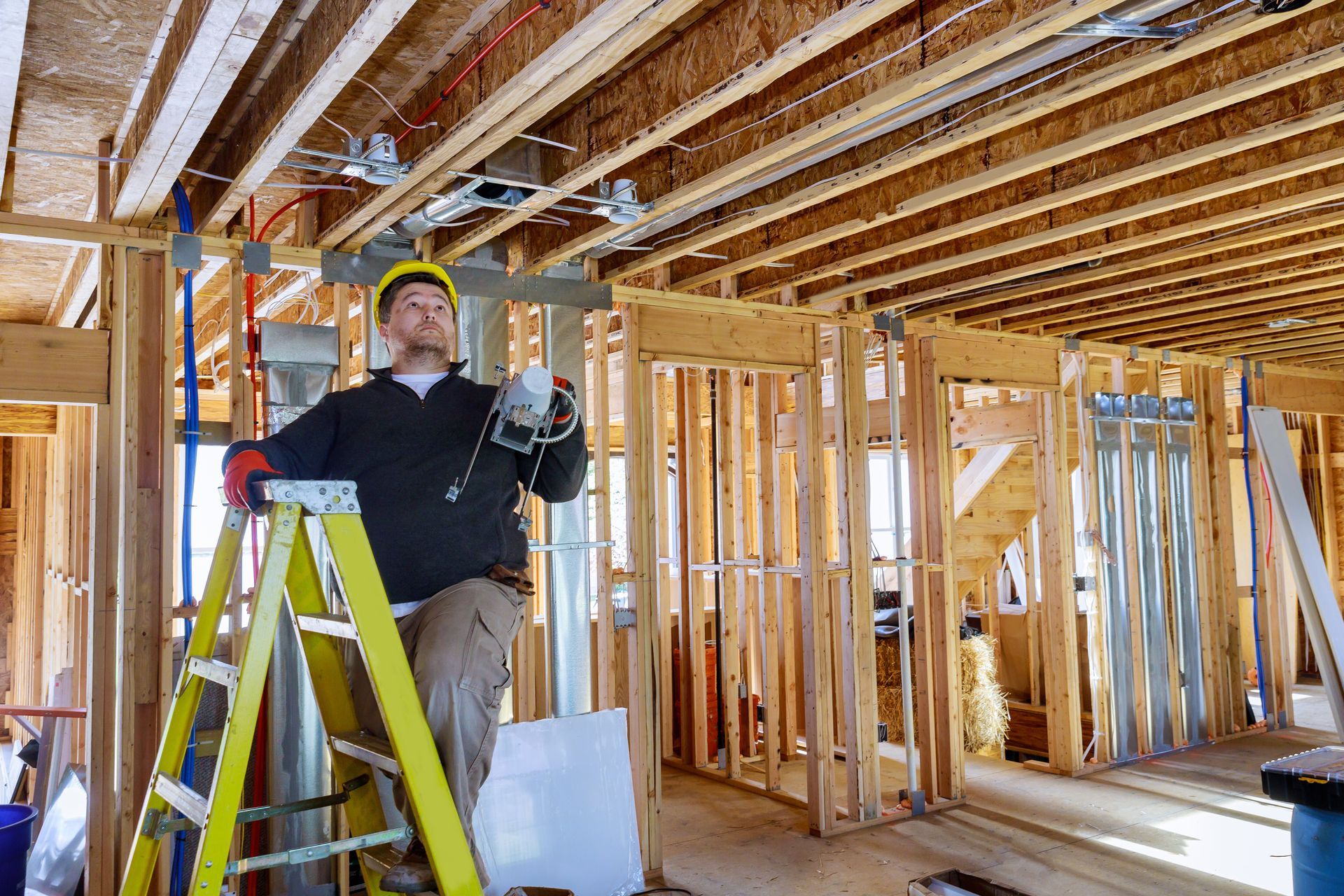 Construction worker on a ladder installs electrical wiring in a wooden framed building.