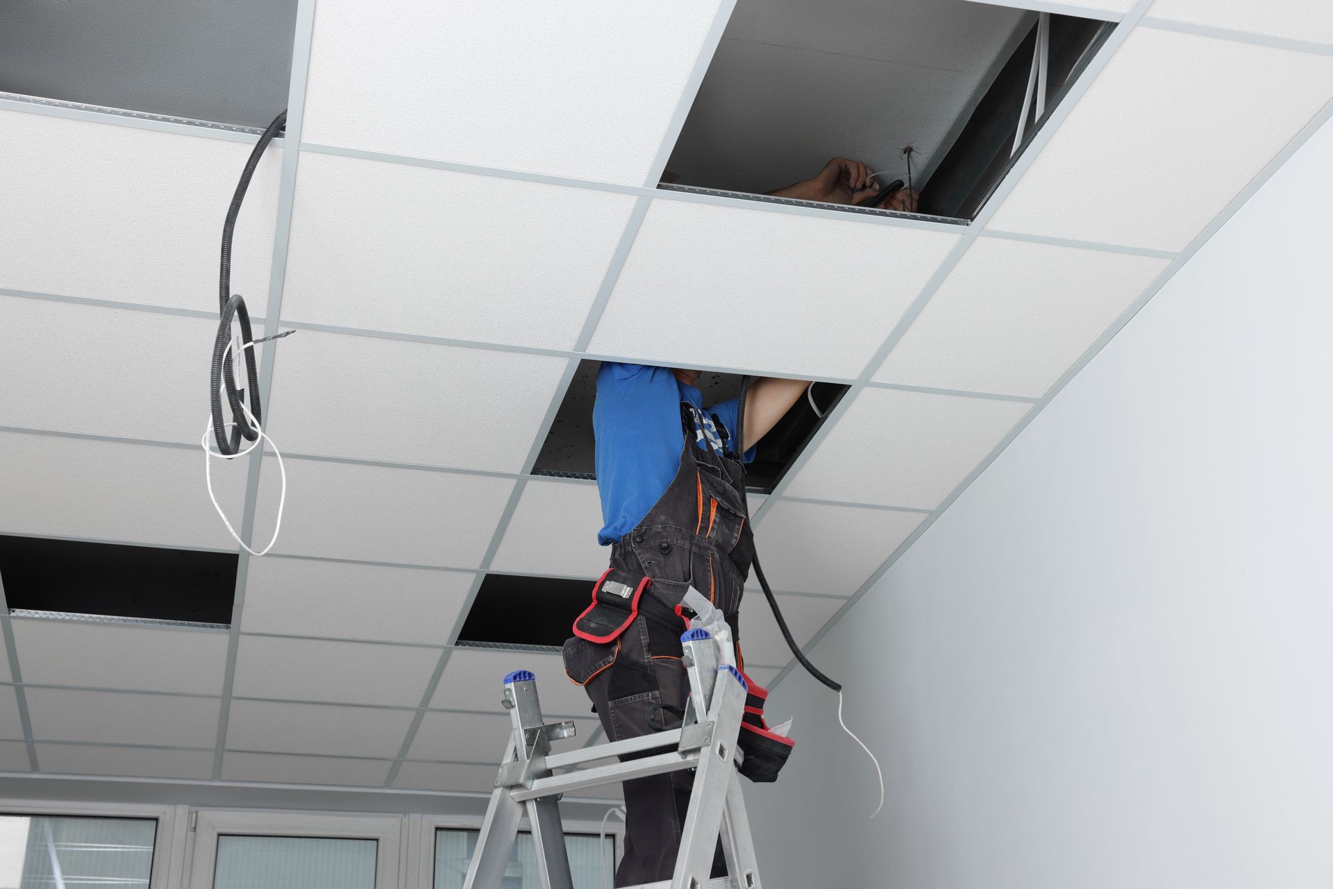 Person on a ladder installing wiring in a drop ceiling with missing tiles.