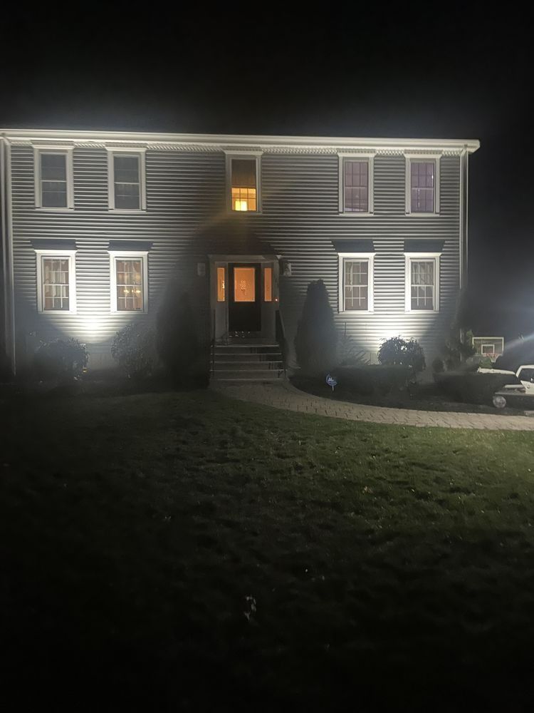Two-story gray house at night with lights on in windows and two front yard spotlights.