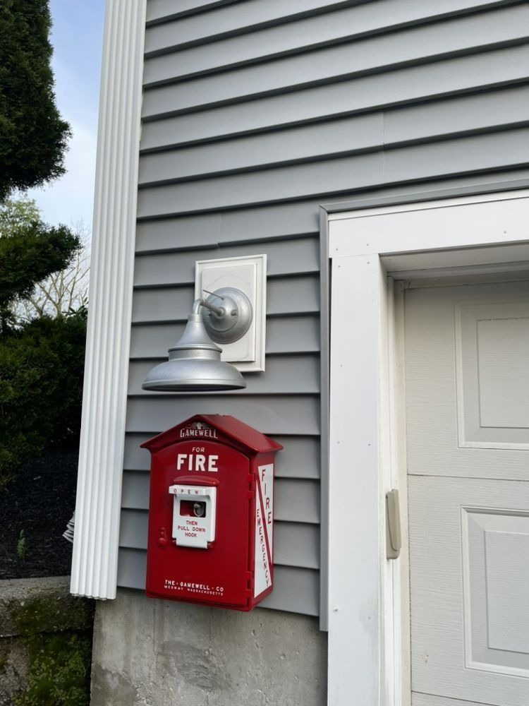 Red fire alarm box and silver lamp mounted on gray siding next to a white door.