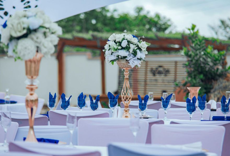 Outdoor wedding reception tables with white chairs, blue napkins, and floral centerpieces under a pavilion