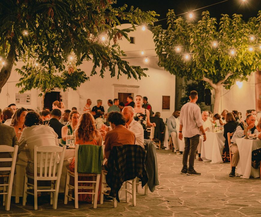 Outdoor evening dinner party under string lights, with guests seated at long tables in a courtyard.