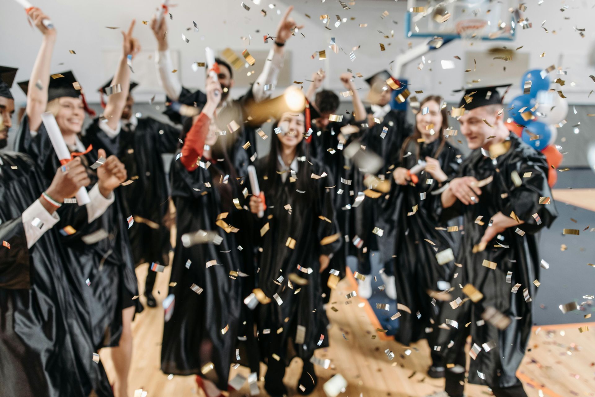 Graduates in black caps and gowns celebrate while confetti falls around them outdoors