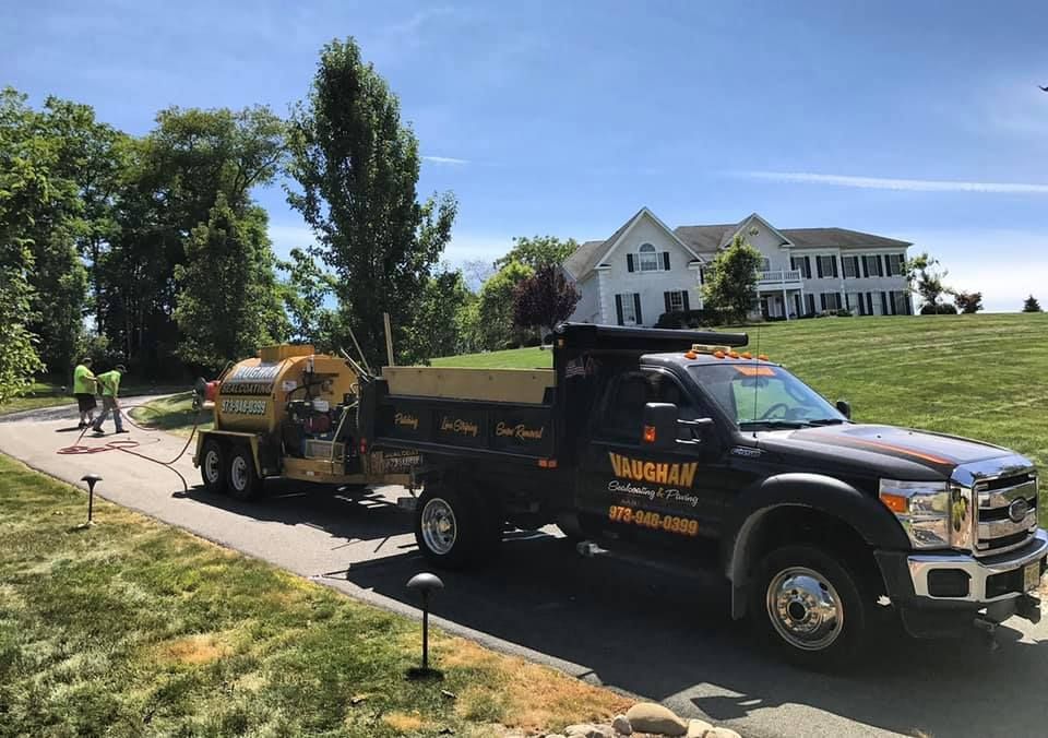 A Vaughan Paving dump truck and equipment trailer parked on a residential driveway near a large two-story house.