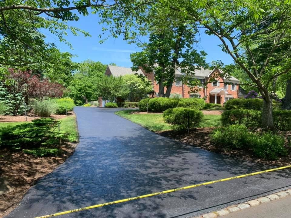 A freshly paved dark asphalt driveway leads toward a brick house surrounded by lush green trees and landscaping.