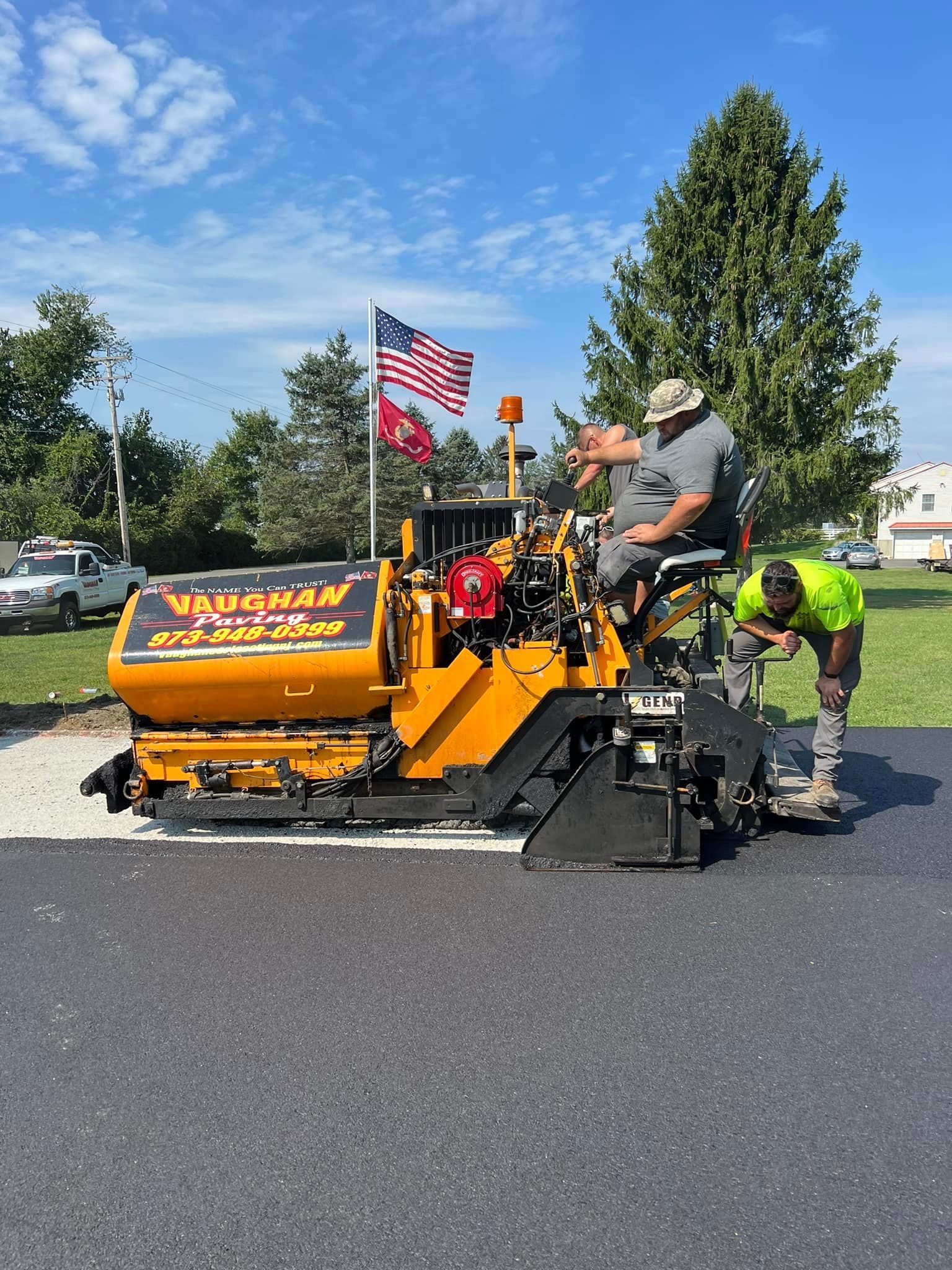 A construction crew operates a yellow asphalt paver on a newly paved driveway under a blue sky with an American flag.