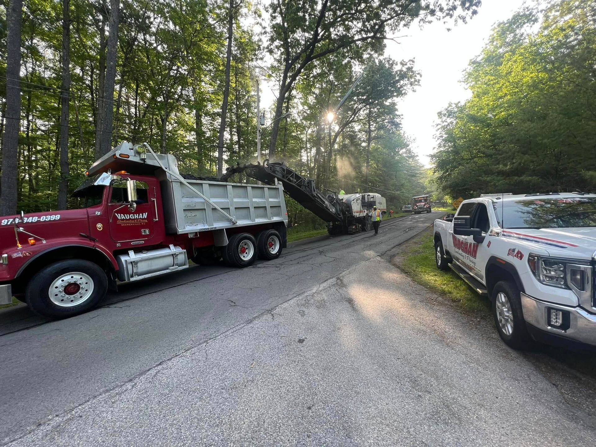 A red dump truck and a white work truck parked on a rural road next to construction equipment clearing asphalt.