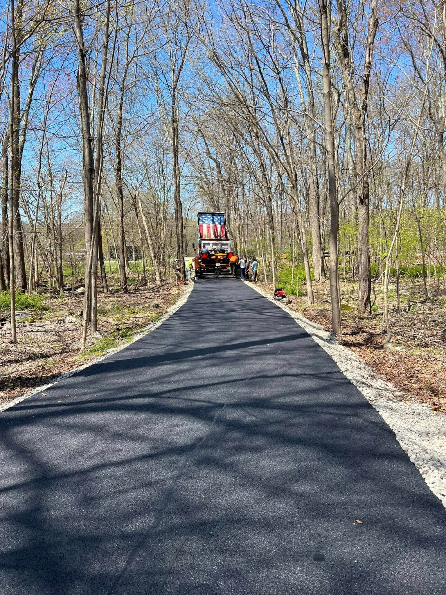 A paved path stretches through a wooded area toward a piece of construction equipment parked in the distance.