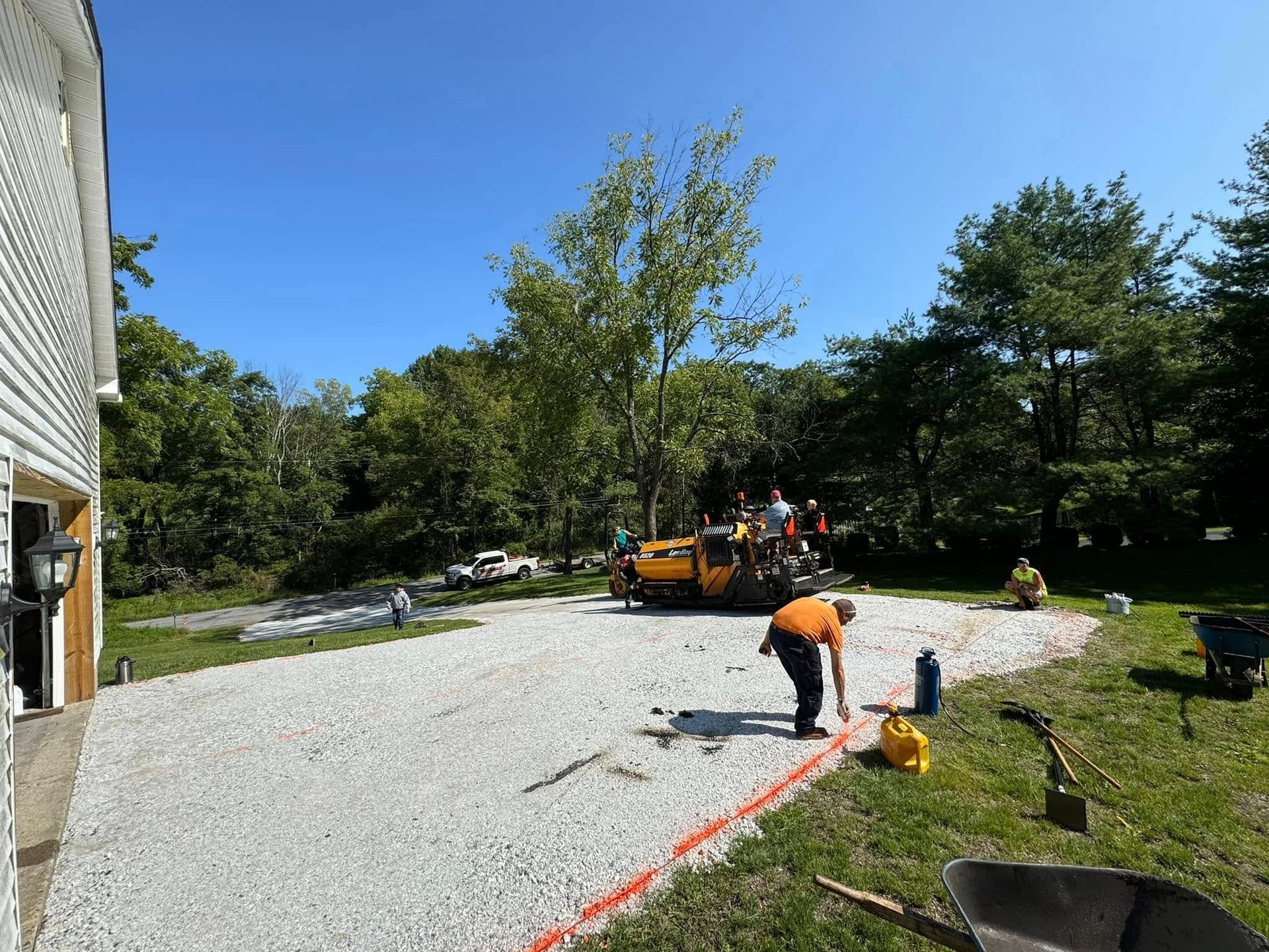 A person in an orange shirt lays out orange caution tape on a gravel driveway, with a construction roller in the back.