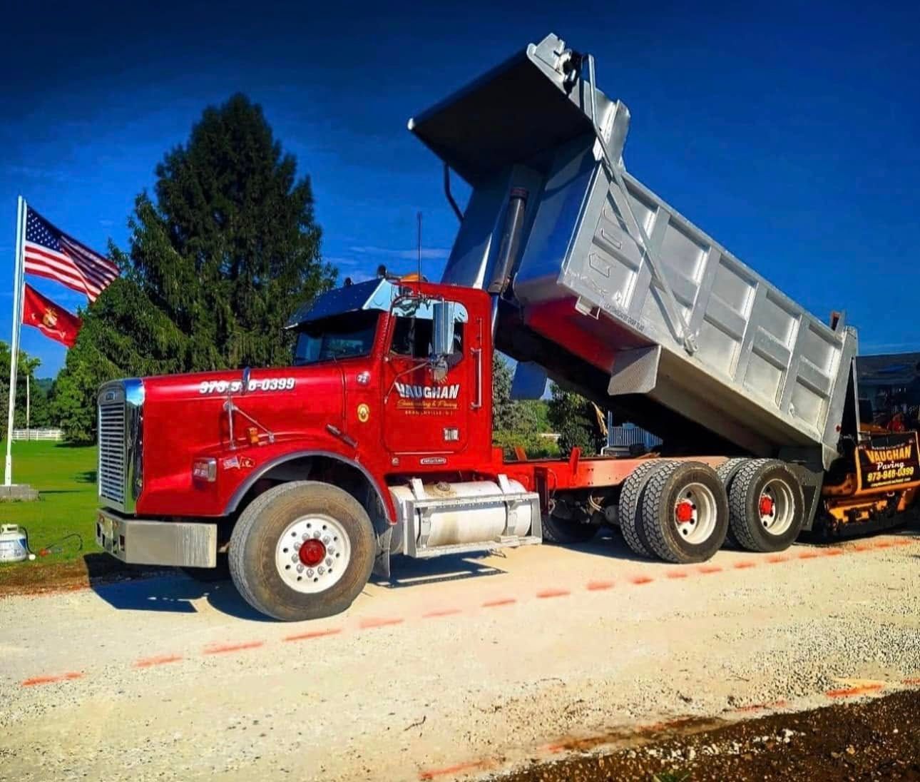A red dump truck with a raised silver trailer parked on a gravel lot next to two American flags.