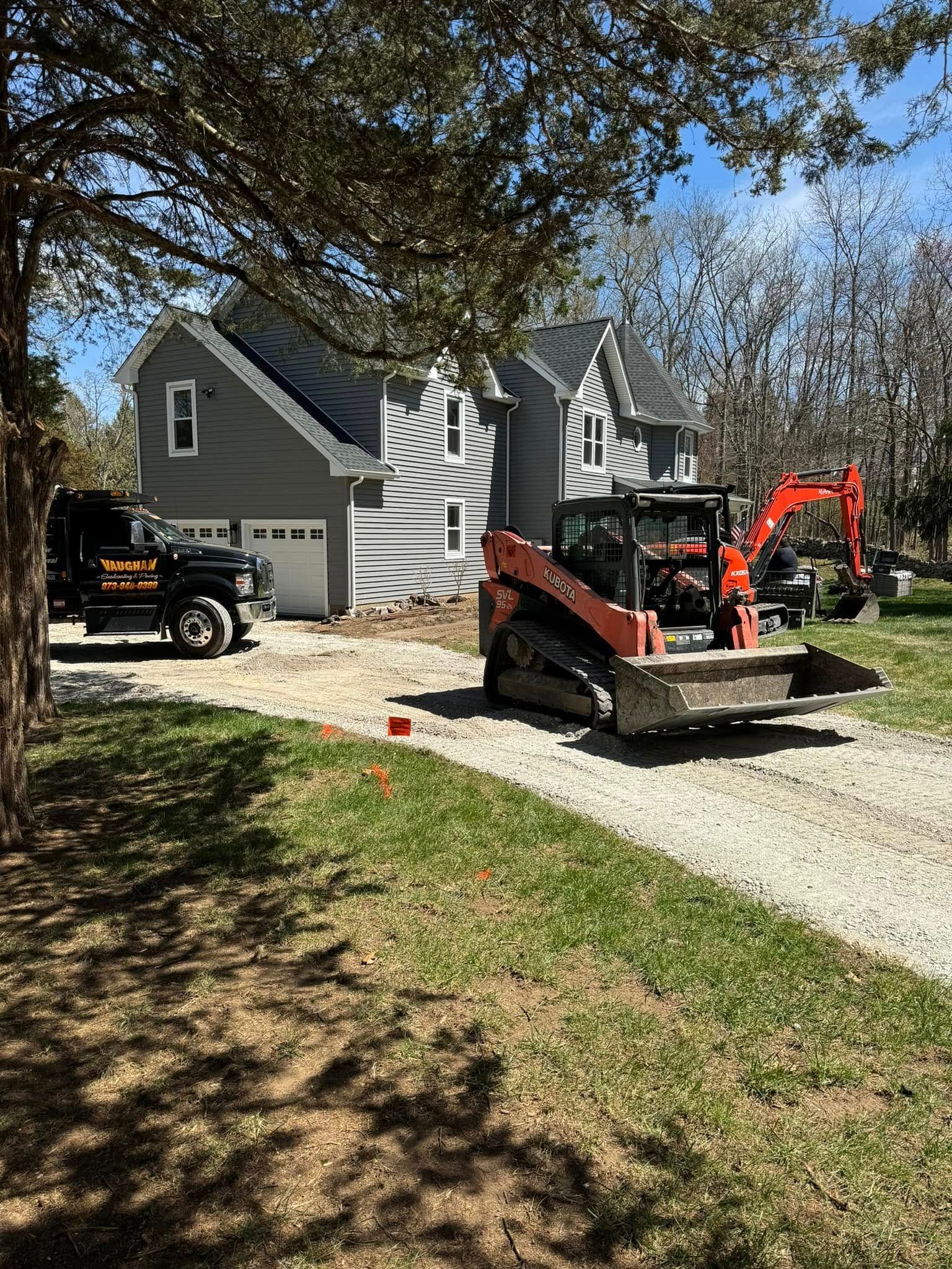 A grey house sits at the end of a gravel driveway with a black utility truck and an orange skid-steer loader parked.