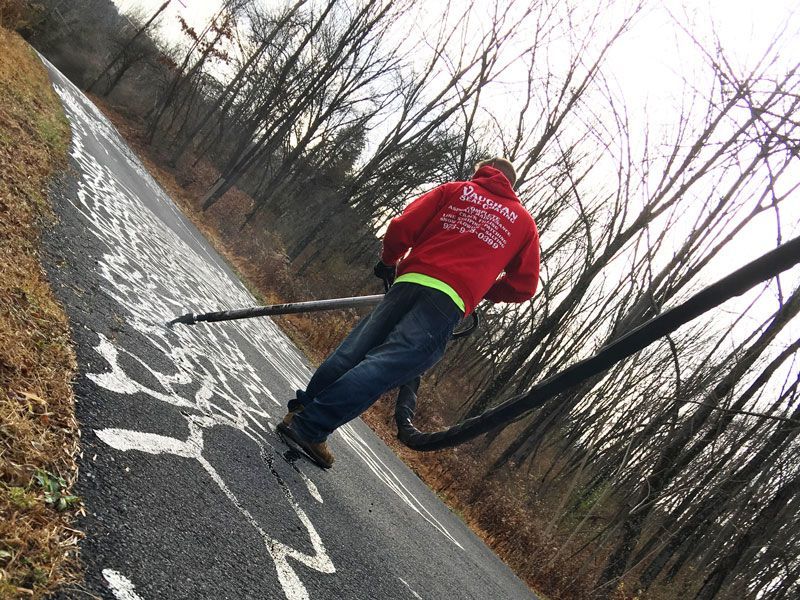 A person in a red hoodie uses a hose to apply white paint to a blacktop road in a wooded area.
