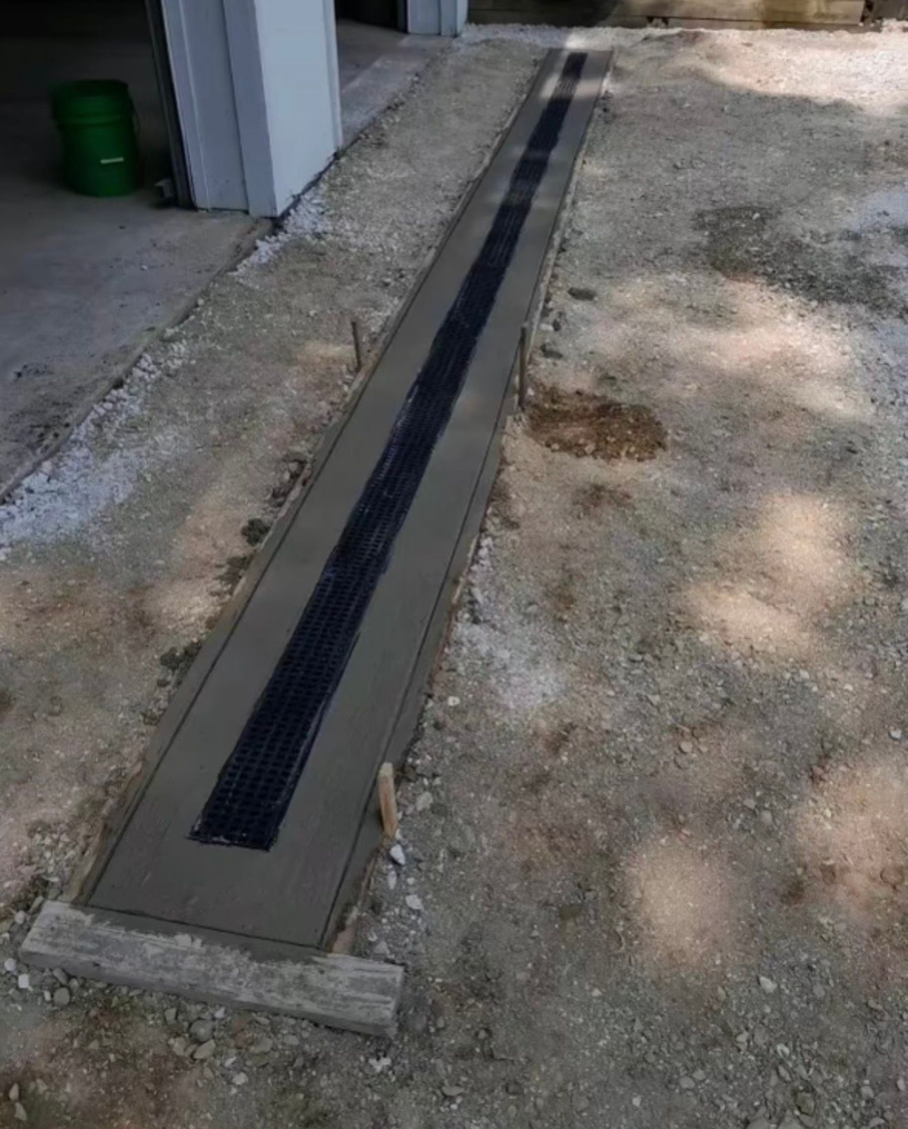 A newly installed black trench drain set into a strip of fresh concrete in a gravel driveway.